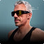 A fit, middle-aged man with short gray hair and a beard wears black sunglasses and a black tank top. A music overlay shows Breathe by The Prodigy is playing. Blue background.