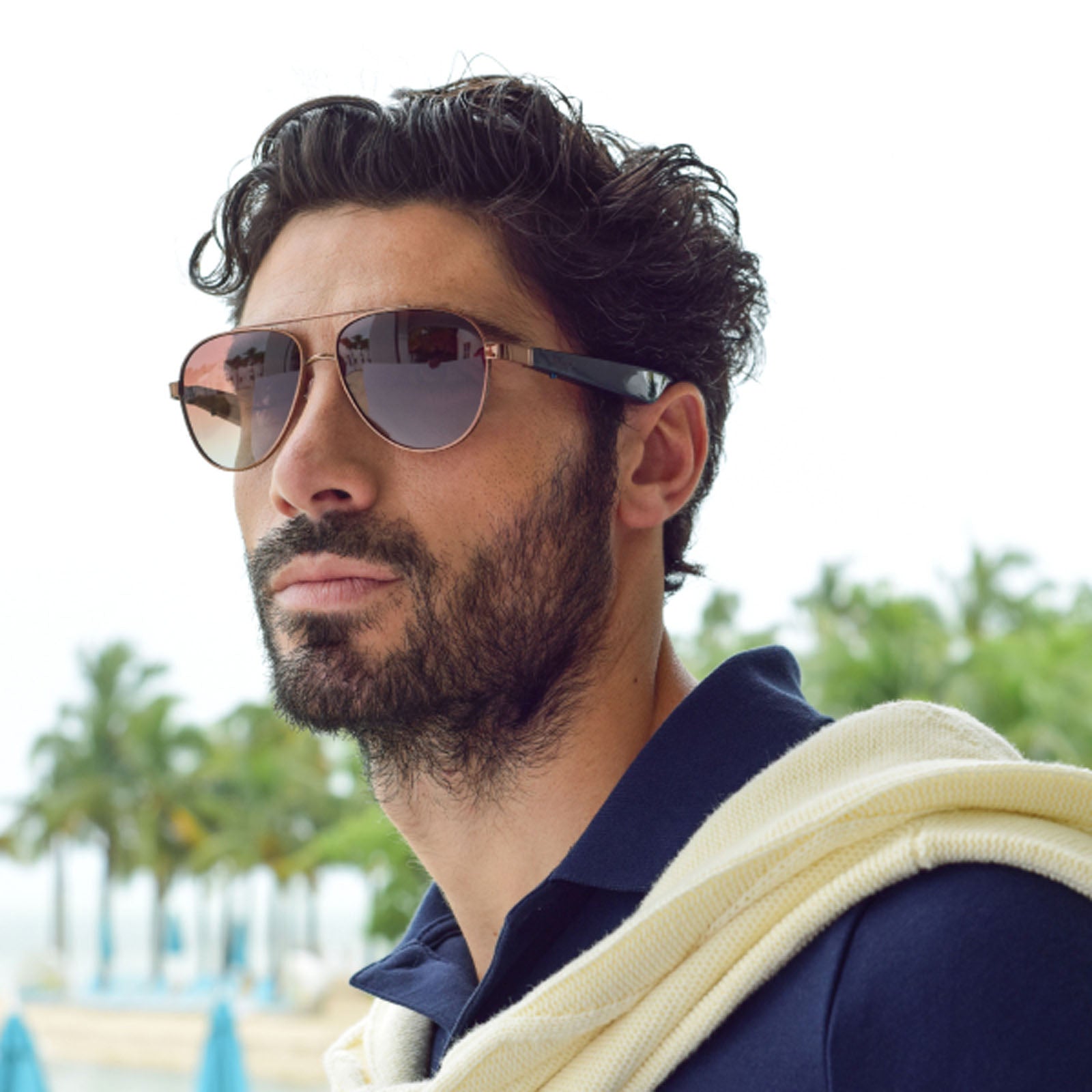 A man with dark curly hair, a beard, and sunglasses—specifically Nautica Admiral polarized aviators—wears a navy polo shirt with a cream sweater over his shoulders, standing outdoors with blurred palm trees and blue umbrellas behind him.