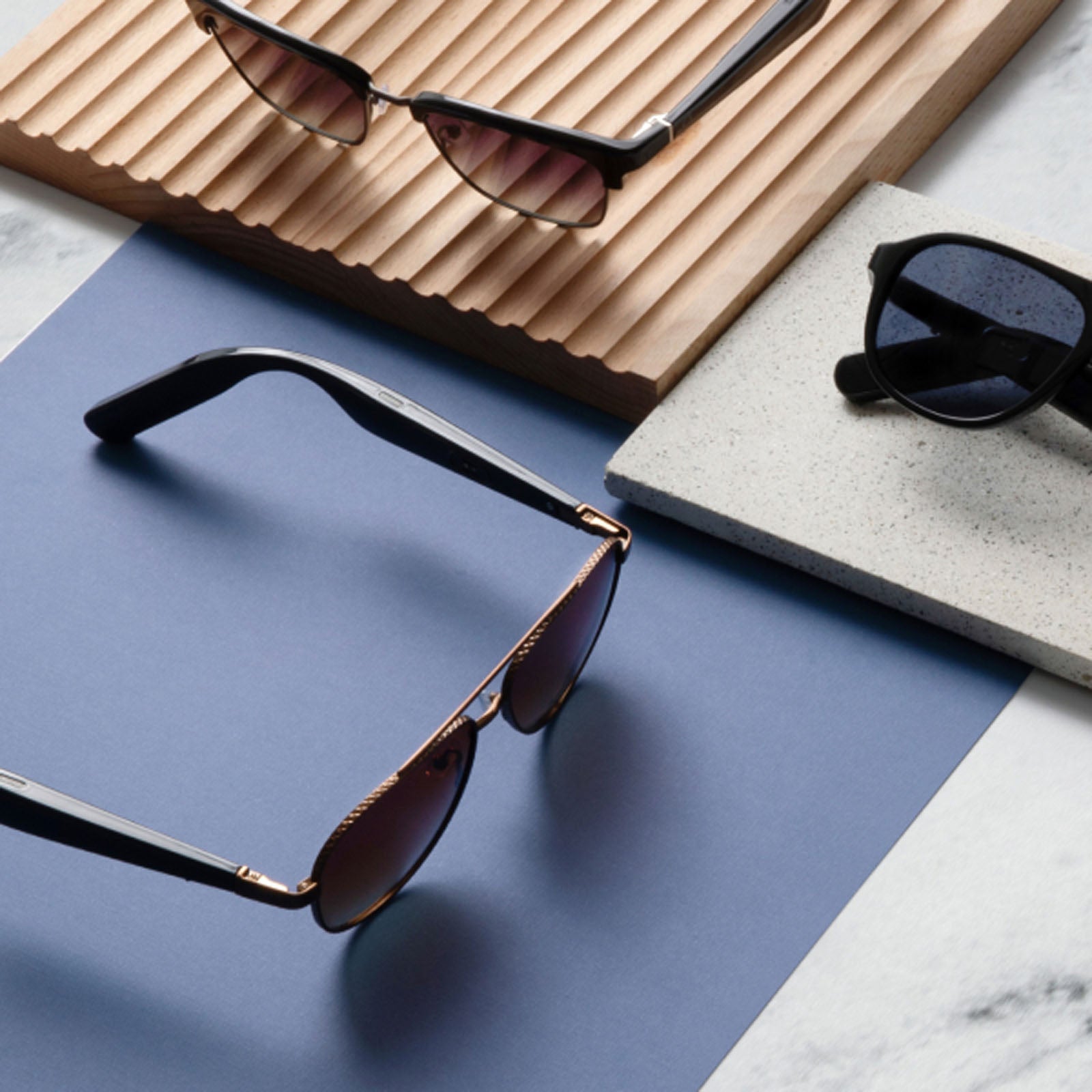 Three pairs of Nautica Admiral smart audio sunglasses are displayed on a blue and white marble surface, accented by wooden and stone trays for a textured, minimalist presentation.