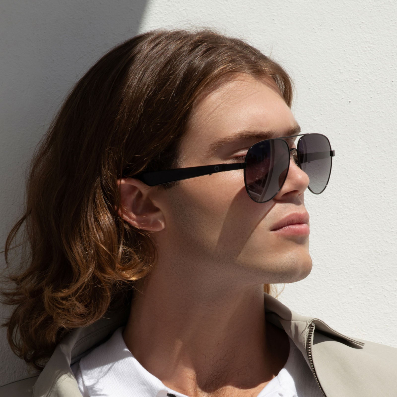 A person with long brown hair wears Lucyd XL’s Antimatter XXL polarized sunglasses and a light jacket, standing against a white wall with sunlight casting a shadow.