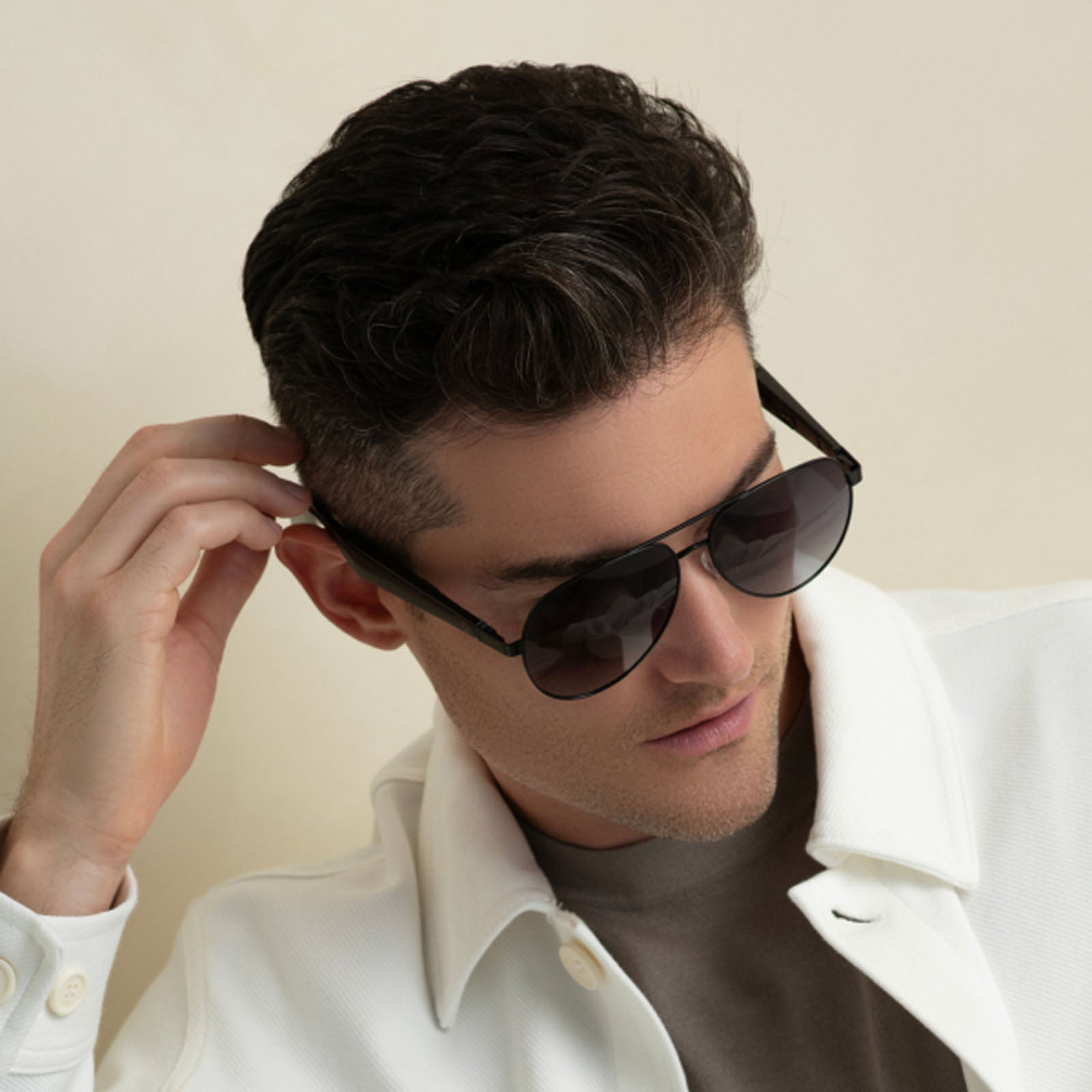 A man with short dark hair wearing Lucyd XL Antimatter XXL polarized sunglasses and a white collared jacket looks slightly downward, adjusting his sunglasses with his right hand against a plain, neutral background.