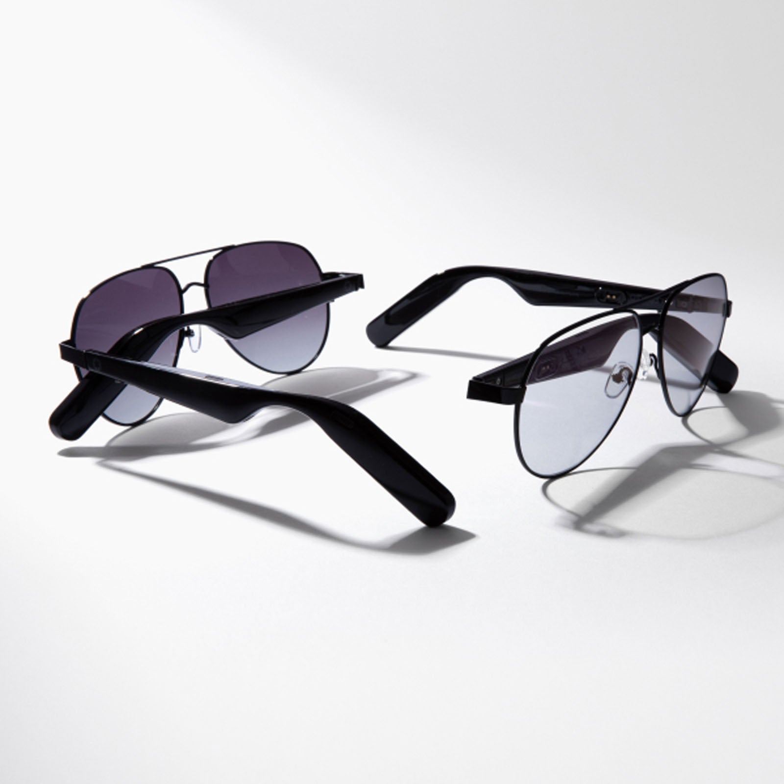 Two pairs of Lucyd XL Antimatter XXL black-framed aviator sunglasses, one with dark lenses and one with lighter, more transparent lenses, sit on a white surface casting soft shadows. Both feature stylish polarized designs.