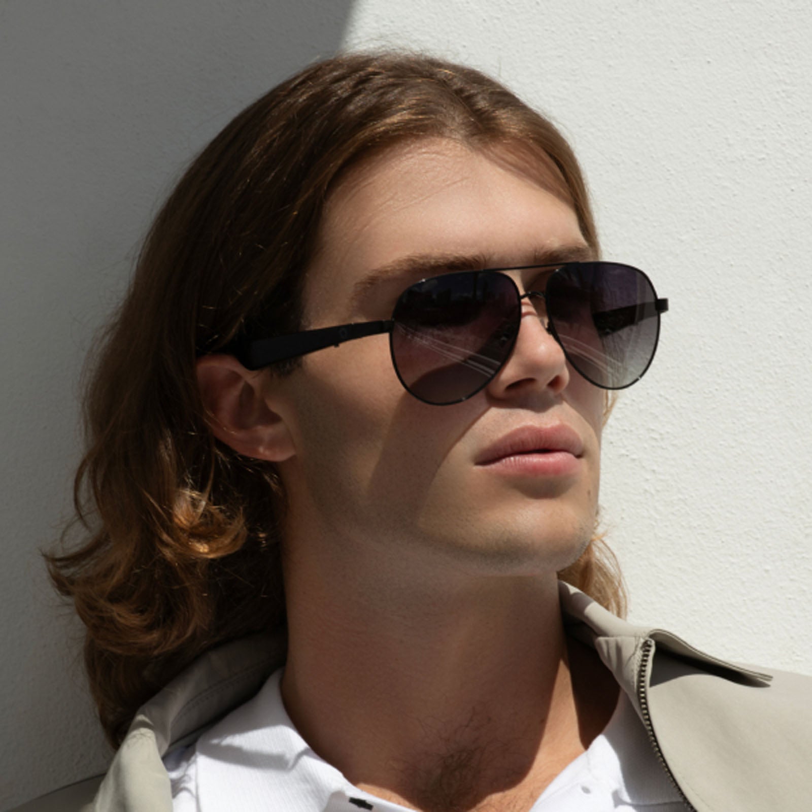 A young man with long light brown hair wears Lucyd XL Antimatter XXL Bluetooth audio sunglasses and a light jacket over a white shirt, standing against a white wall in partial sunlight.