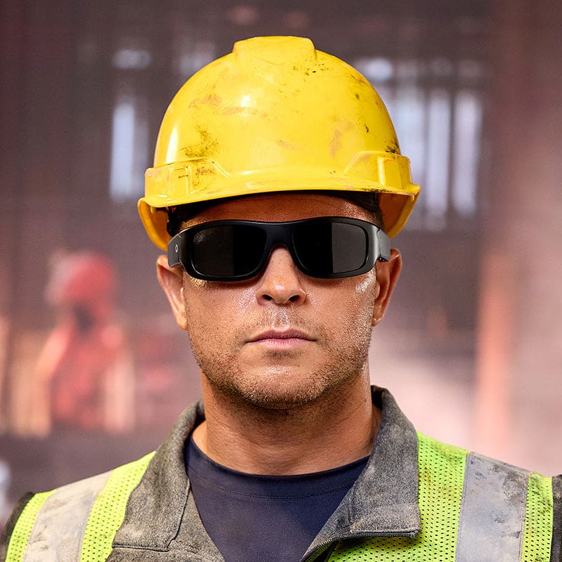 A construction worker in a yellow hard hat, high-visibility vest, and Lucyd Armor | Black Polarized safety glasses stands before a blurred industrial background.