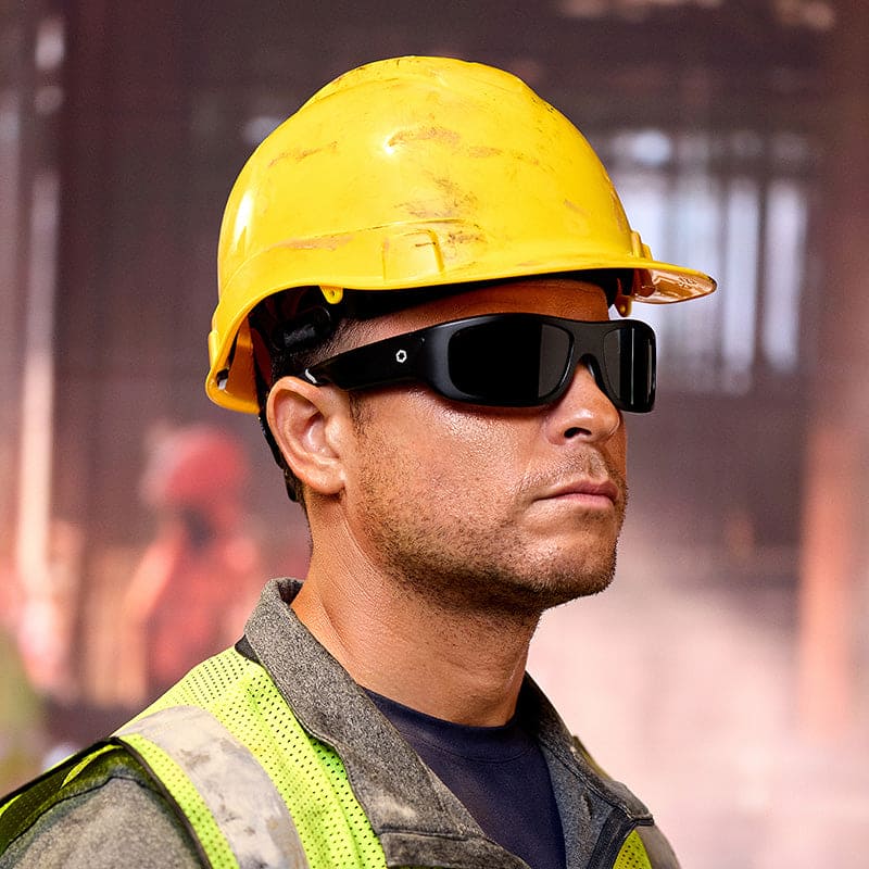 A construction worker in a yellow hard hat, Lucyd Armor | Black Polarized sunglasses by Lucyd, and a reflective safety vest stands indoors with a blurred industrial background and another worker visible in the distance.