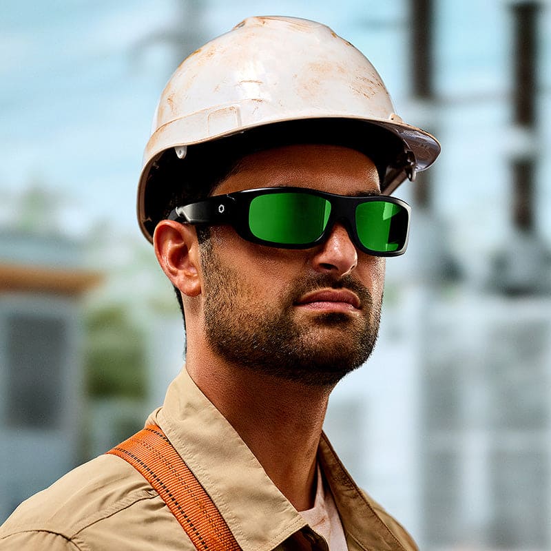 A man stands outdoors at a construction site wearing a white hard hat, beige work shirt, and Lucyd Armor | Green Mirror Polarized safety glasses.