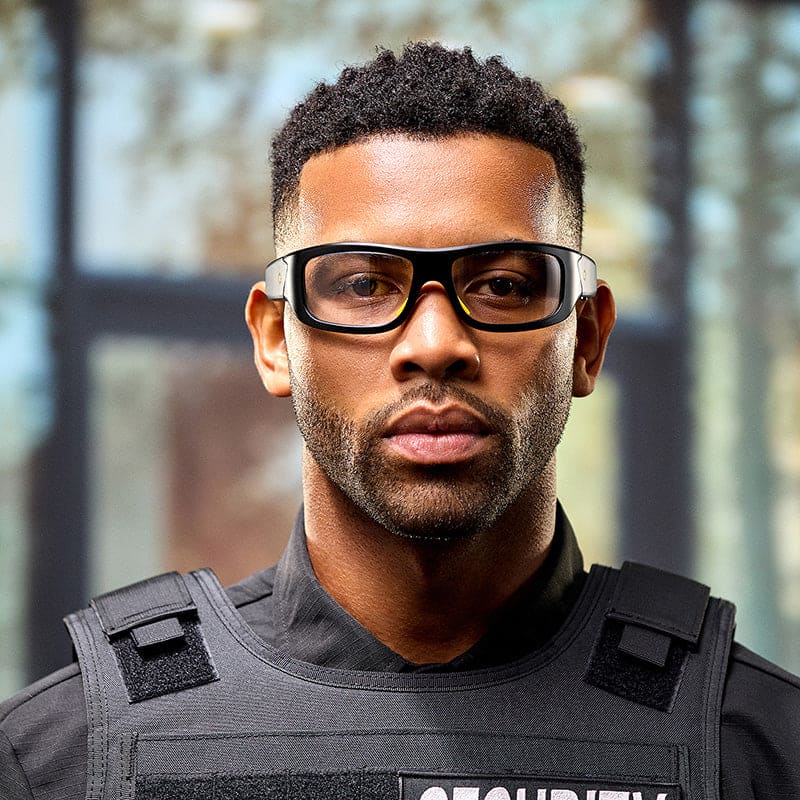A man wears Lucyd Armor Z87.1 safety glasses and a black security vest indoors, looking directly at the camera with a serious expression. The background is blurred.