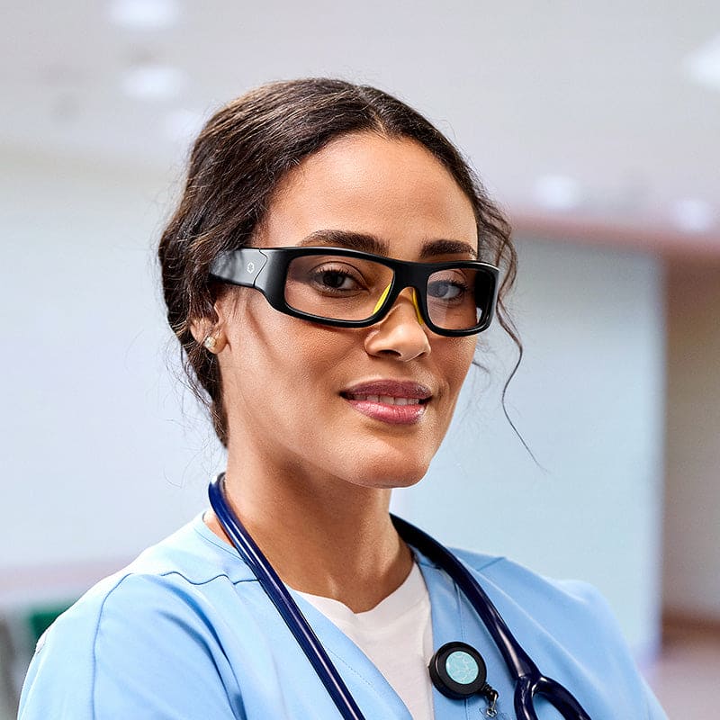 A woman in blue medical scrubs and a stethoscope smiles in a modern healthcare setting while wearing Lucyd Armor | Slim Photochromic Bluetooth audio glasses.