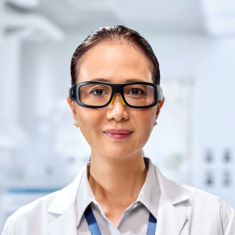 A woman wearing Lucyd Armor | Slim Photochromic glasses and a white lab coat smiles softly while standing in a bright laboratory setting.