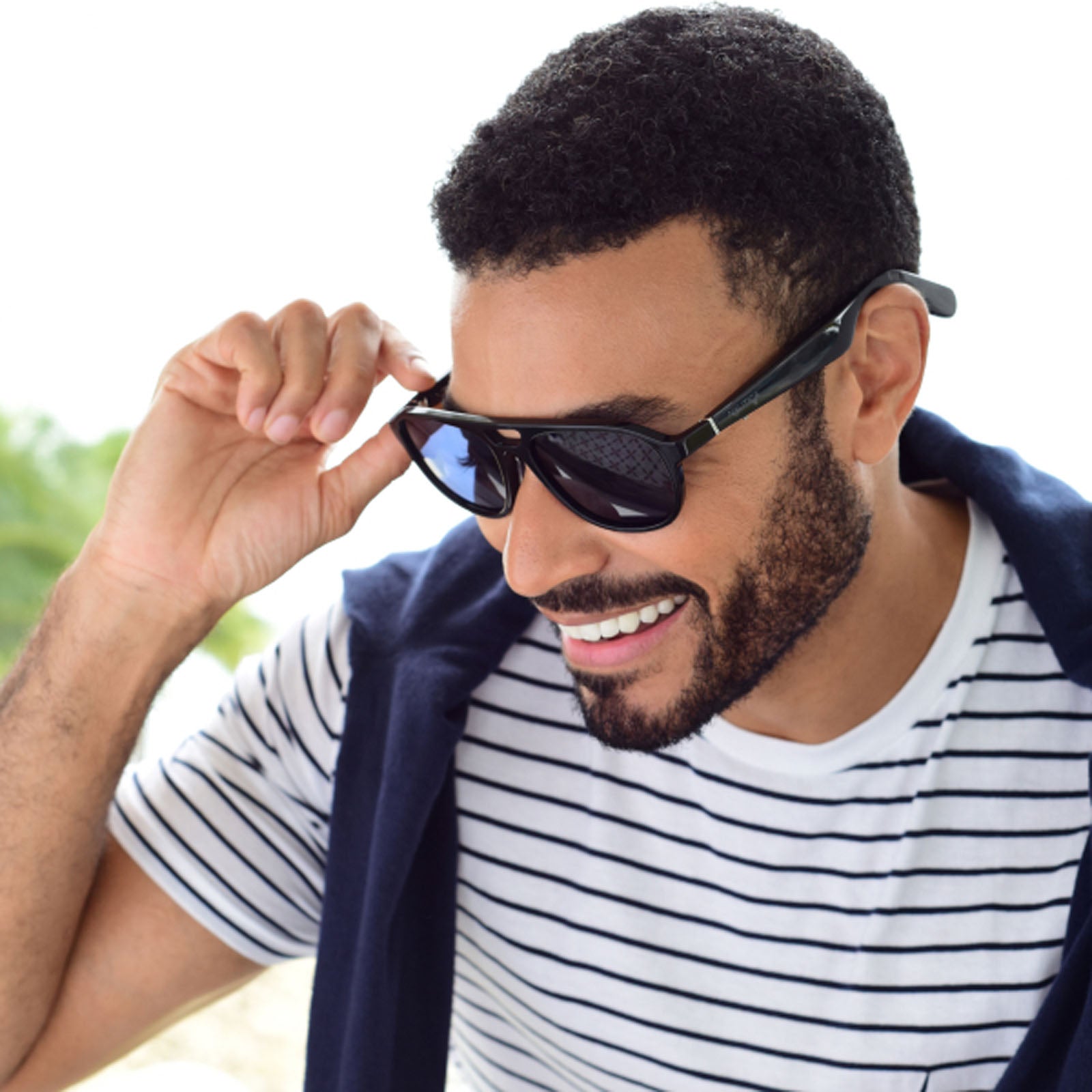 A smiling bearded man wearing Nautica Commander polarized aviator sunglasses and a striped shirt with a navy sweater over his shoulders holds his sunglasses while looking slightly downward.