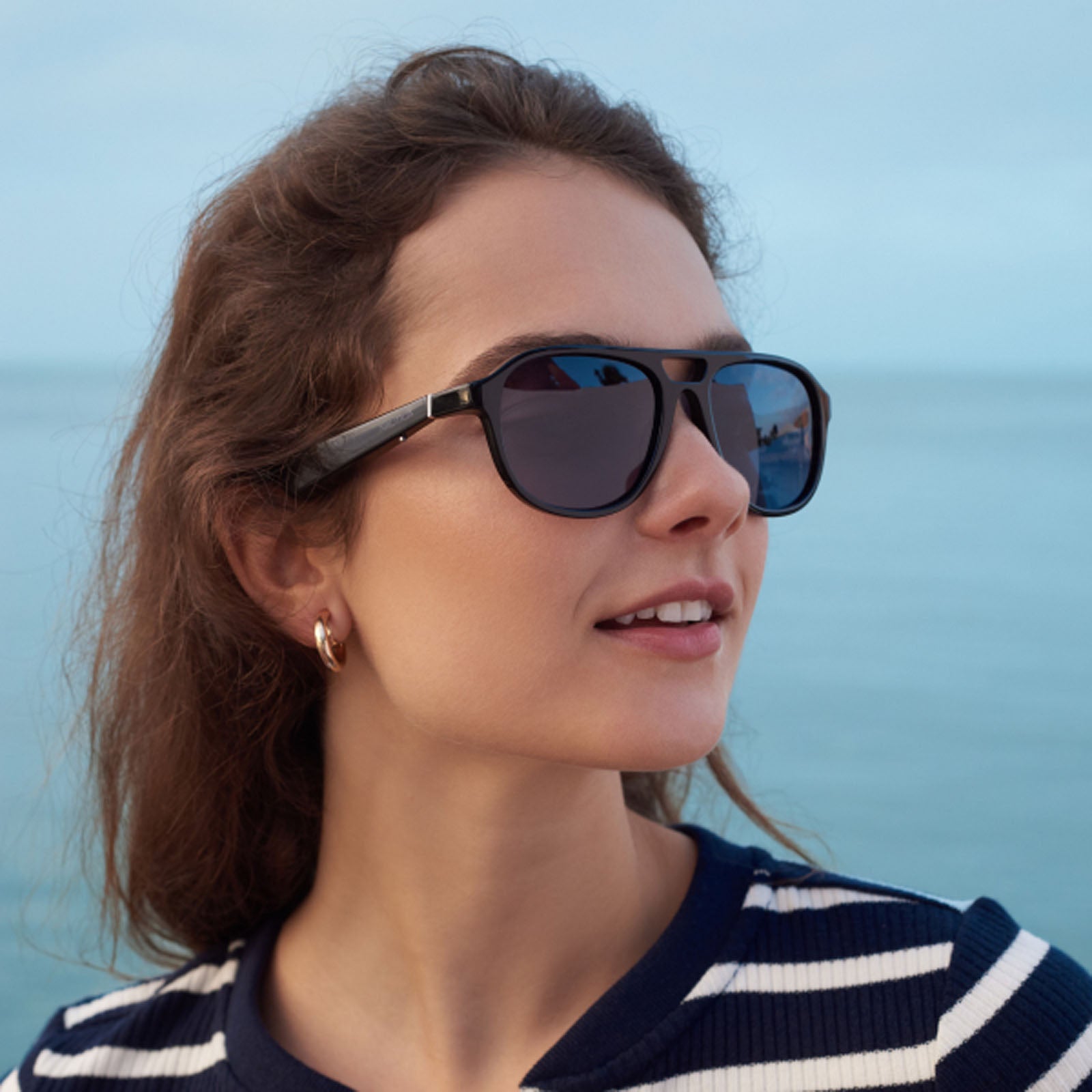 A woman with wavy brown hair wears Nautica Commander polarized aviator sunglasses and a navy-and-white striped shirt, standing outdoors by the sea under a clear sky.