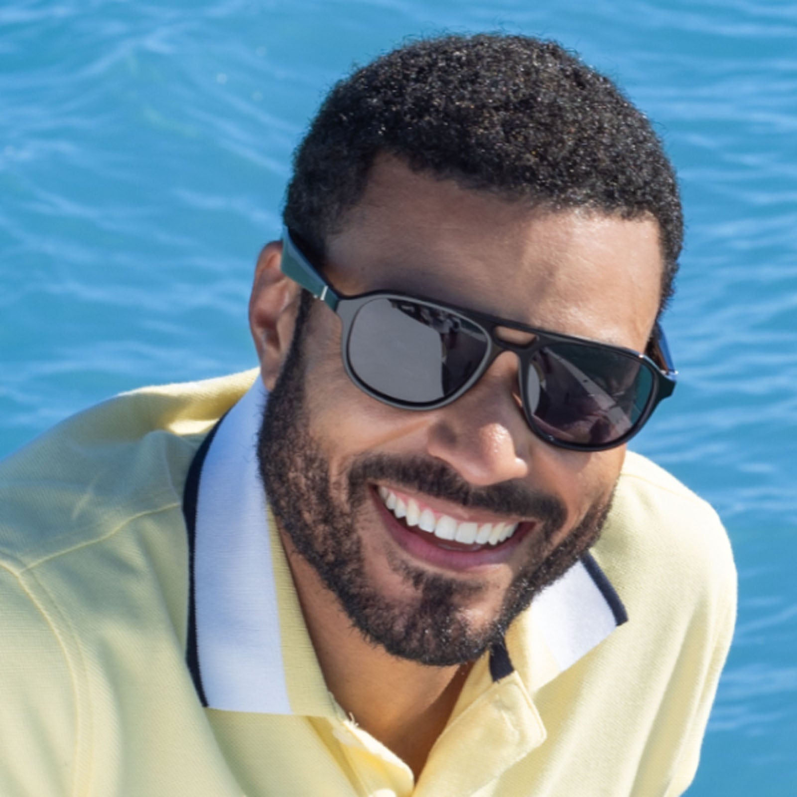 A man with short curly hair, a beard, and Bluetooth sunglasses smiles in a yellow Nautica Commander polo shirt, outdoors with bright blue water in the background.