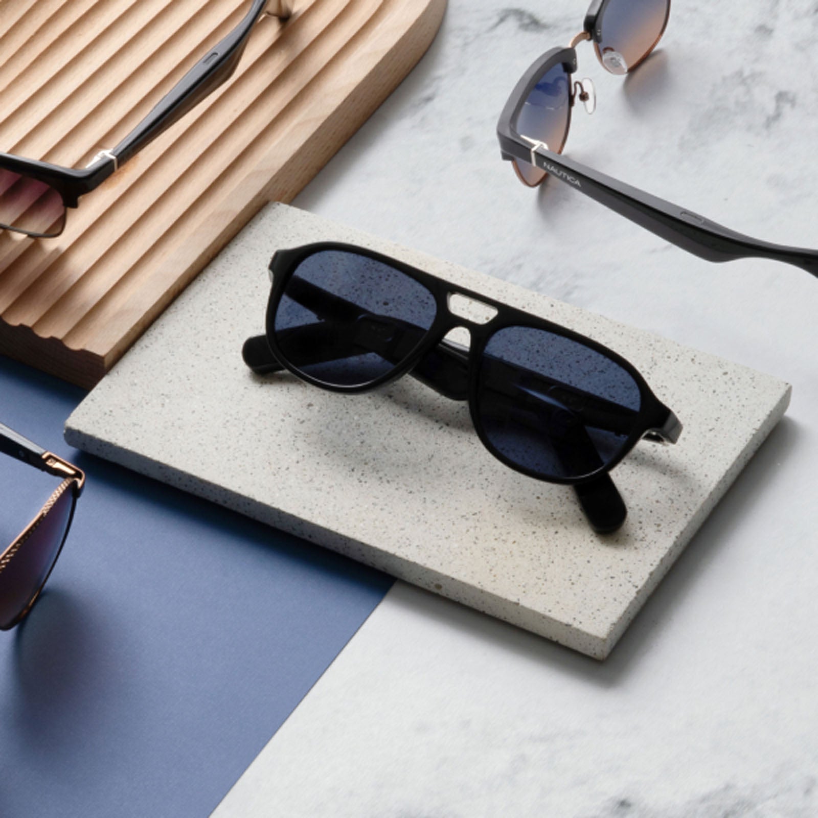 The Nautica Commander black Bluetooth sunglasses are showcased on a rectangular stone slab, with three other pairs of sunglasses arranged nearby on a marble surface featuring blue and wooden accents.