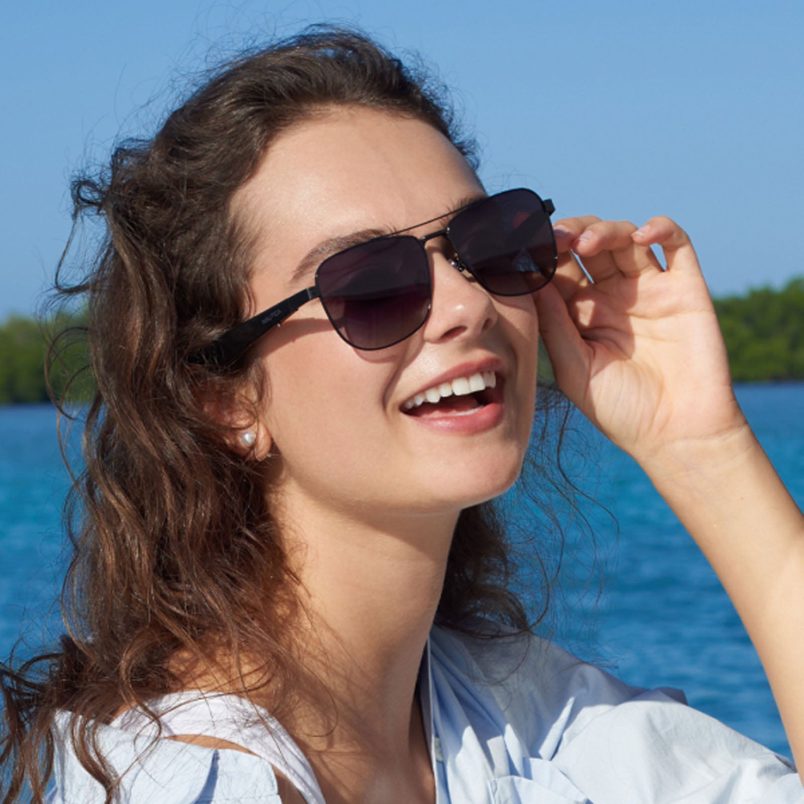 A woman with wavy brown hair smiles while wearing Nautica Corsair polarized aviator sunglasses near blue water, with green trees and a clear sky in the background.