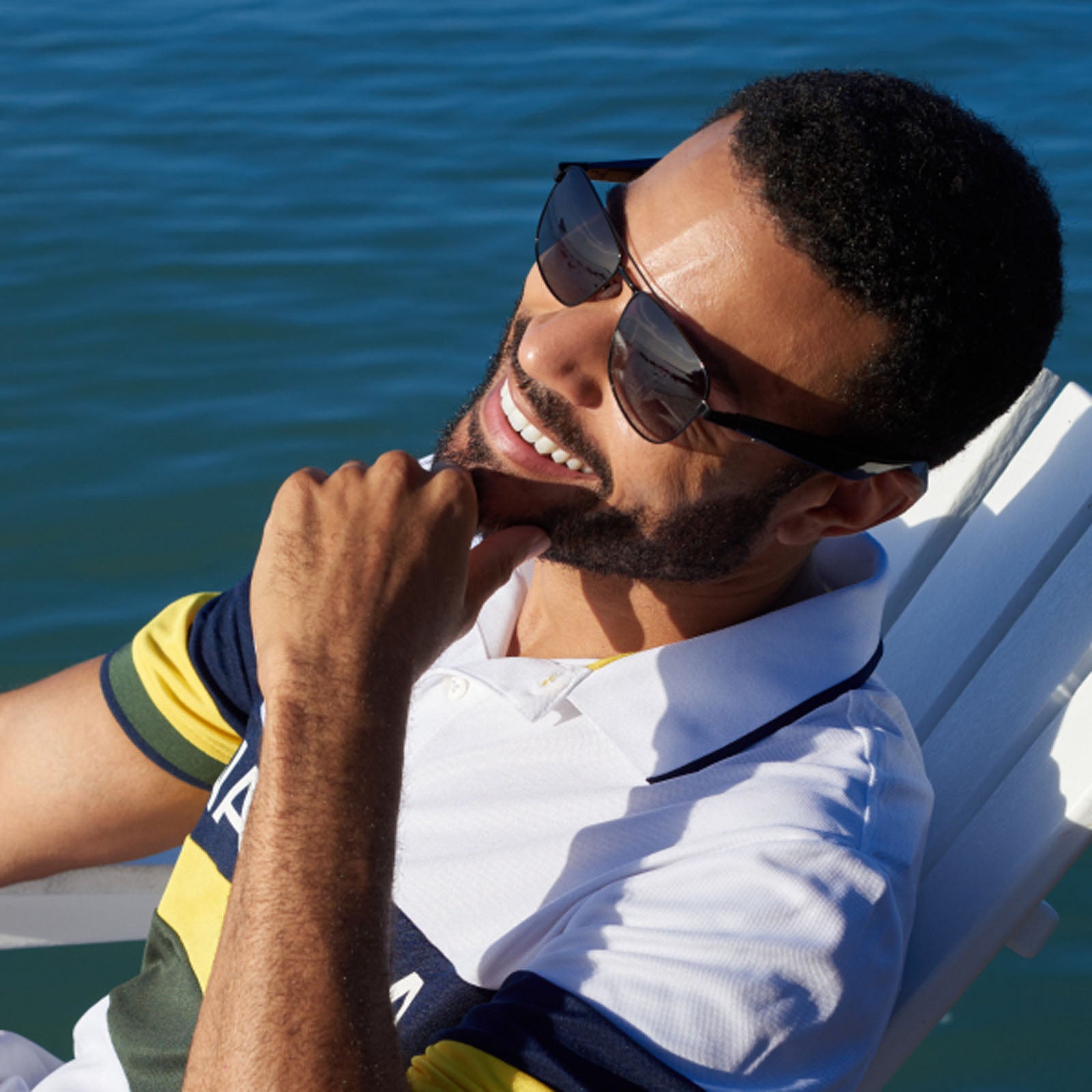 A man in Nautica's Corsair smart sunglasses and a striped polo shirt smiles as he relaxes on a white chair by the water in the bright sun.
