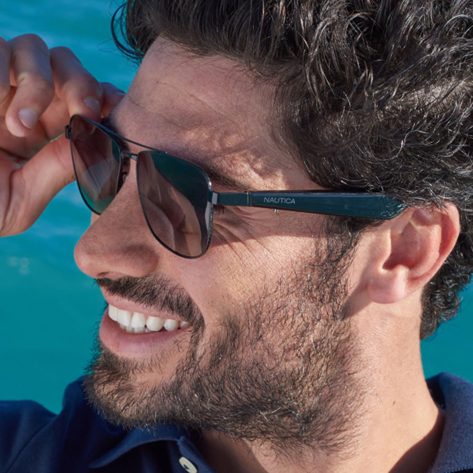 A man with curly dark hair and a beard smiles, wearing Nautica Corsair sunglasses and a navy blue shirt, holding one arm of his glasses as blue water shimmers behind him.