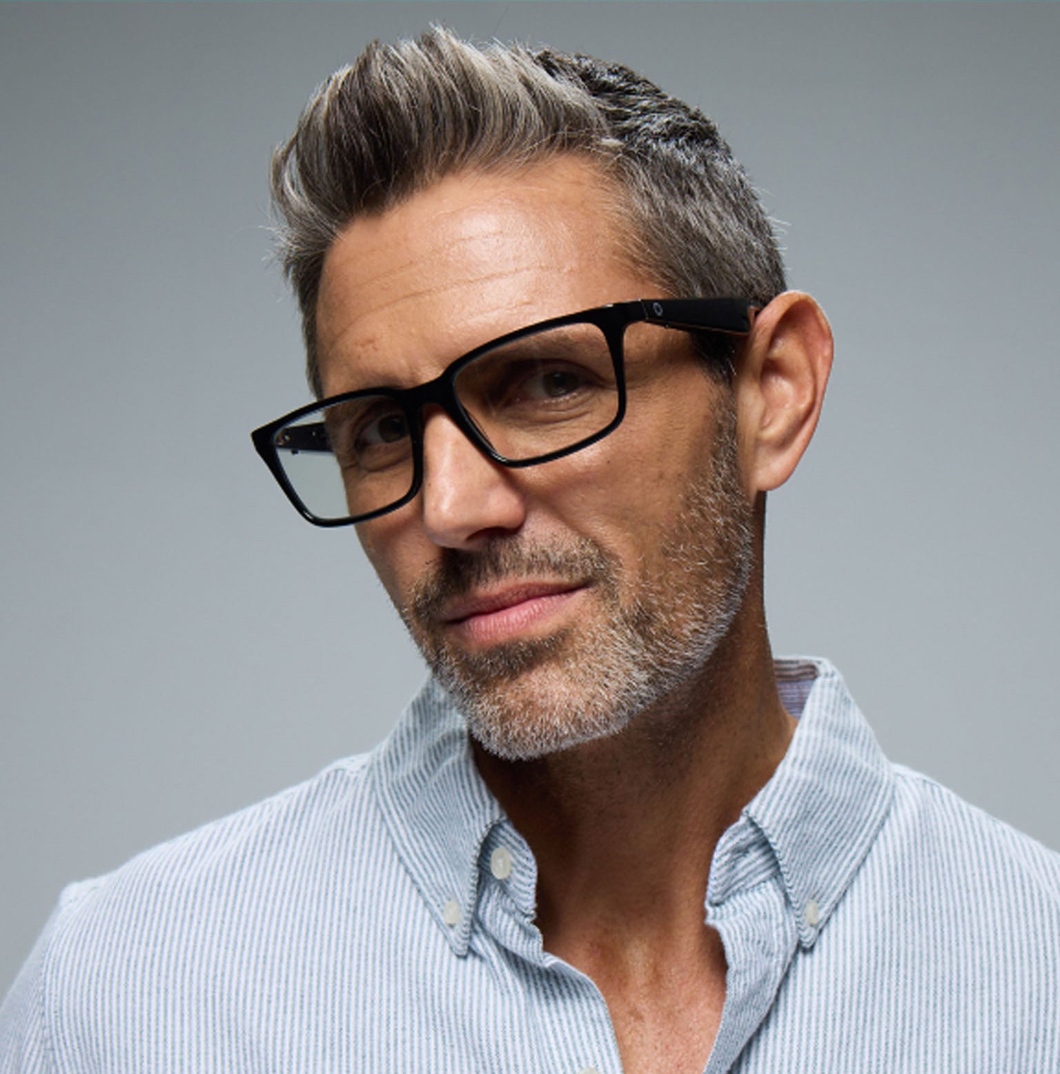 A middle-aged man with salt-and-pepper hair and stubble wears Lucyd Darkside black-rimmed Bluetooth audio sunglasses and a light blue striped shirt, looking confidently at the camera against a plain background.
