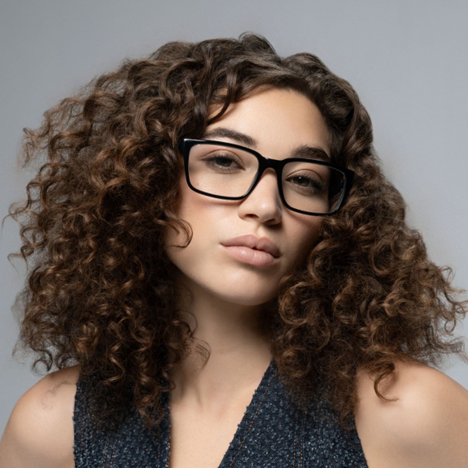 A young woman with curly brown hair wears Lucyd’s Darkside black rectangular Bluetooth audio sunglasses, looking at the camera with a neutral expression. She pairs them with a sleeveless textured top against a plain gray background.