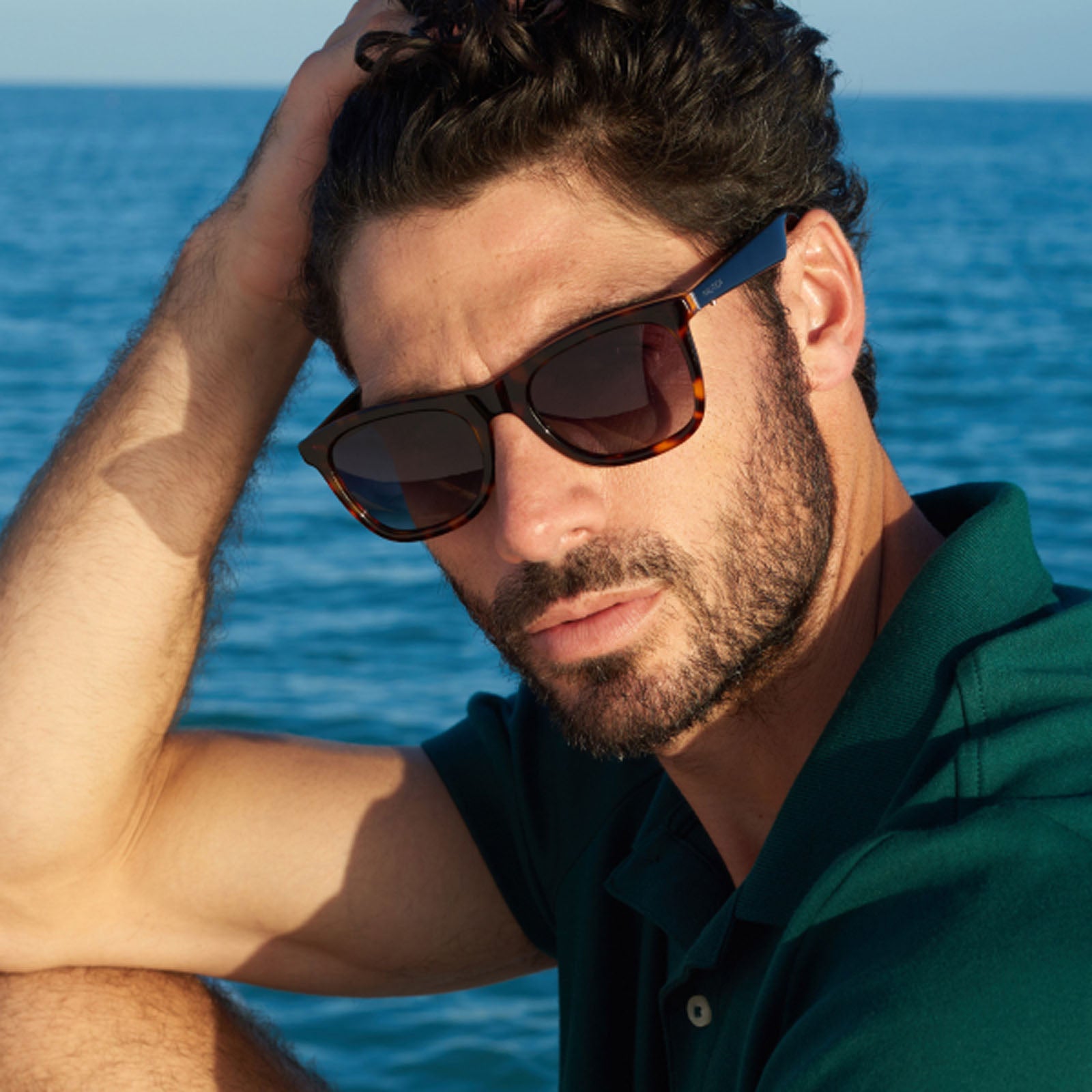 A man with dark hair and a beard wears Nautica’s Dockside smart sunglasses and a dark green polo shirt, sitting by the sea with one hand on his head, looking at the camera against an ocean and blue sky backdrop.