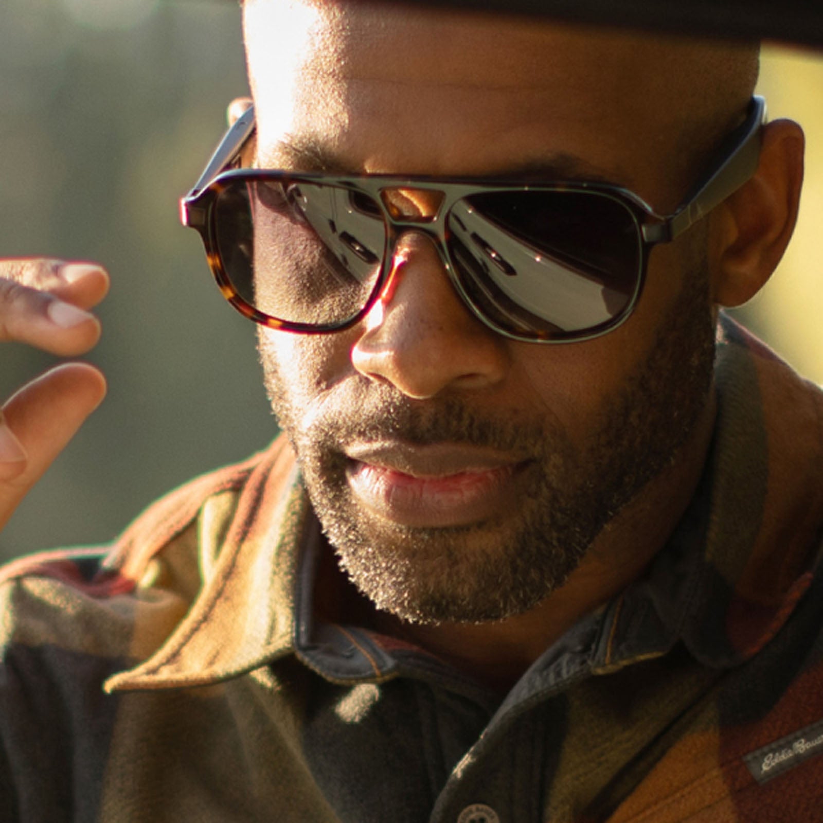 A man with a trimmed beard wears oversized, dark Eddie Bauer Explorer smart glasses with polarized UV400 lenses and a plaid shirt, raising his left hand slightly. Sunlight highlights his face and glasses; the background is softly blurred.