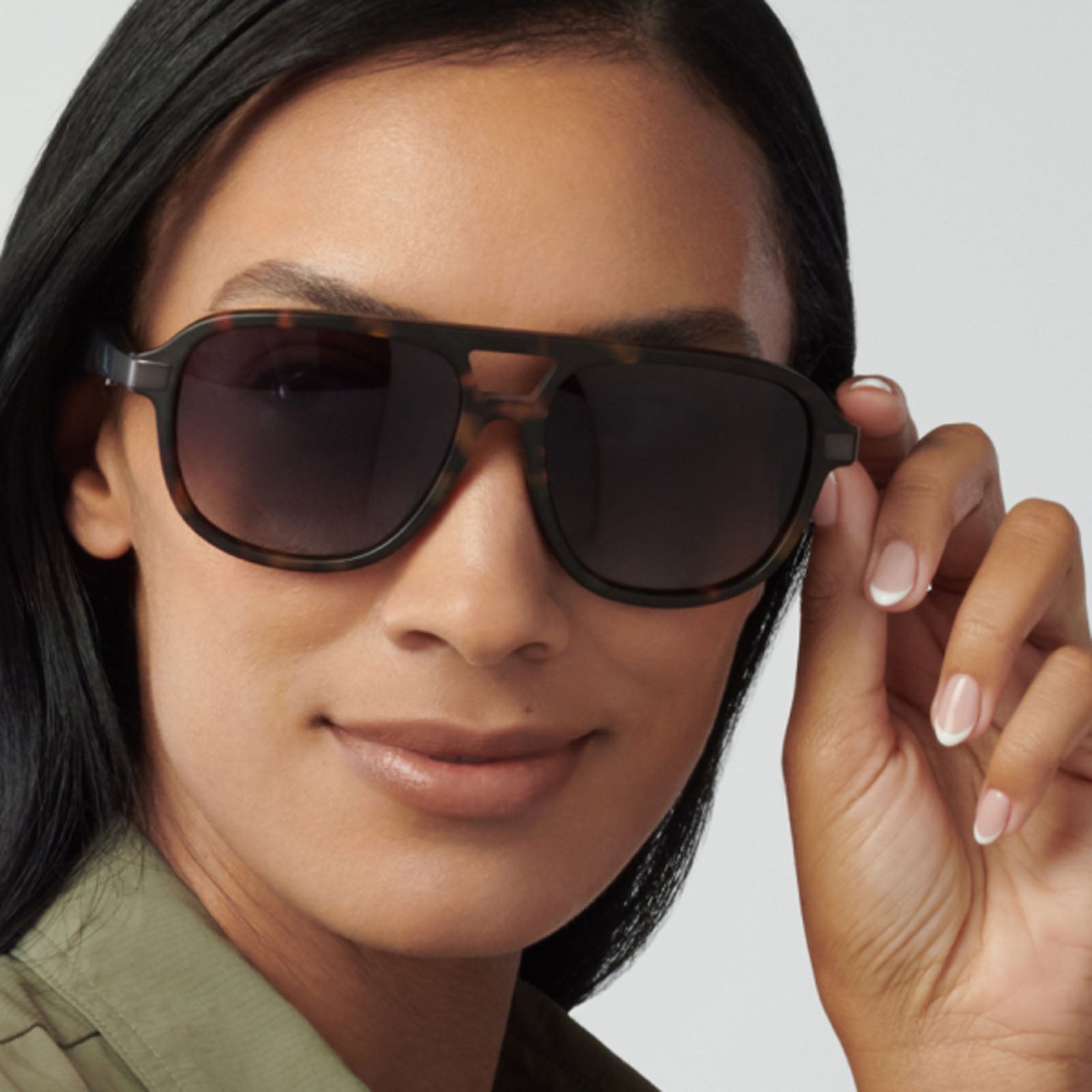 A woman with long dark hair smiles slightly while wearing Eddie Bauer Explorer sunglasses, featuring large polarized UV400 lenses, and a green top. She holds the frame of the sunglasses near her face with one hand.