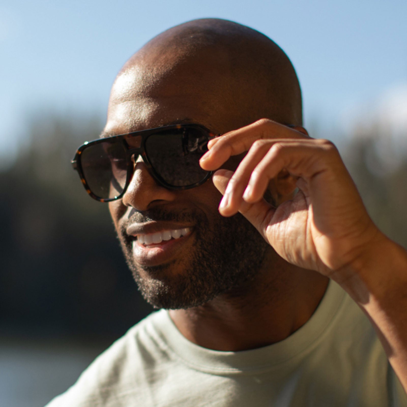 A smiling, bearded bald man wearing large, dark Eddie Bauer Explorer smart glasses and a light shirt stands outside in bright sunlight, holding one side of his Bluetooth sunglasses.