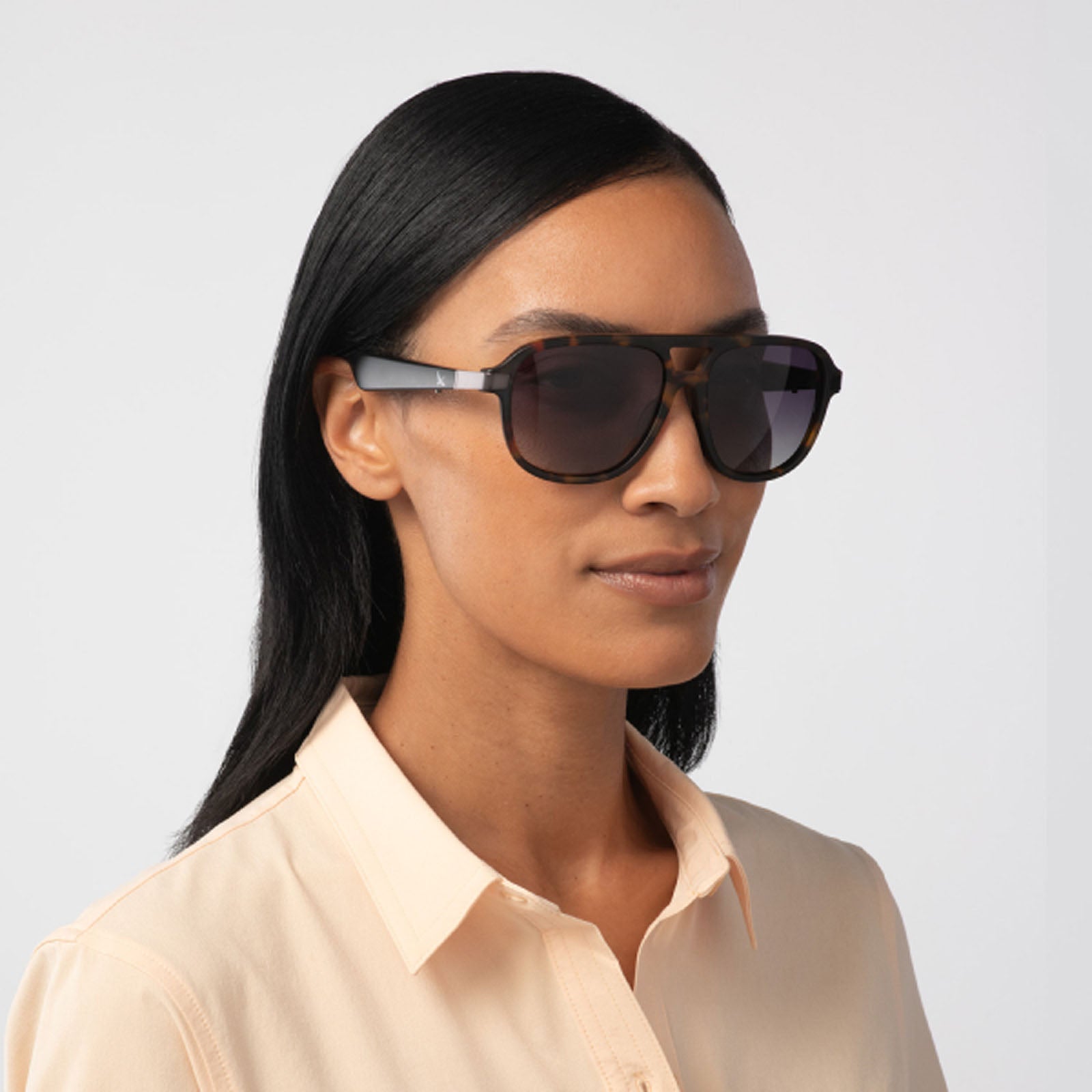 A woman with straight black hair models Eddie Bauer's Explorer sunglasses—large, polarized UV400 lenses—paired with a light peach collared shirt against a plain light background.