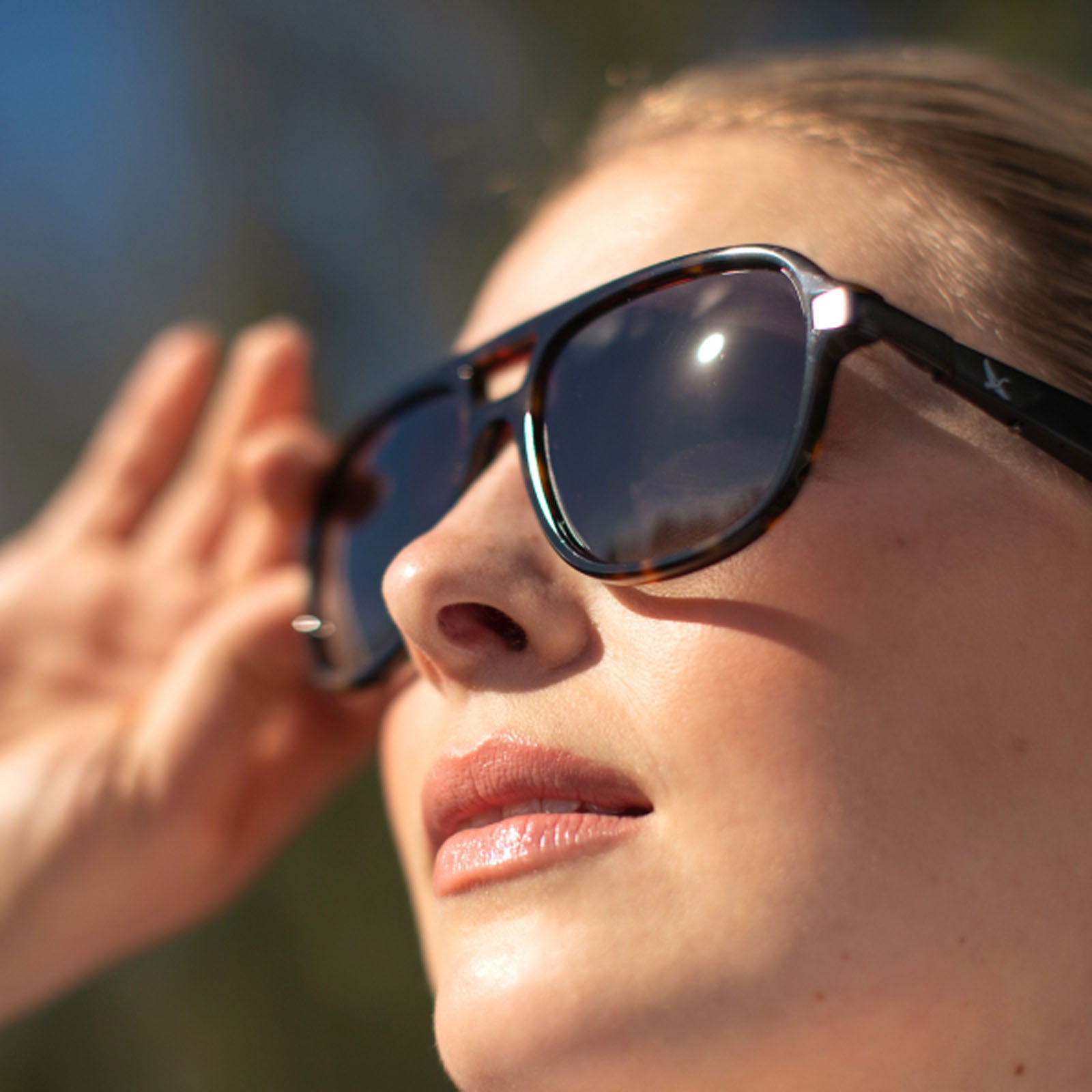 Close-up of a person wearing Eddie Bauer Explorer sunglasses with large, dark, polarized UV400 lenses reflecting sunlight. Their lips are slightly parted and one hand is raised near their face.