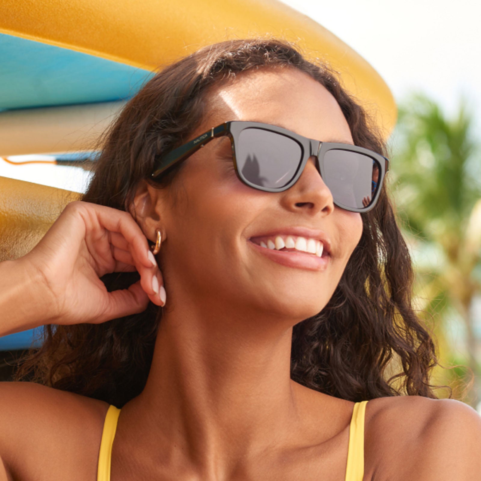 A smiling woman with curly brown hair wears Nautica Fathom UV400 sunglasses and a yellow swimsuit, standing outdoors by colorful surfboards with palm trees and a sunny sky in the background.