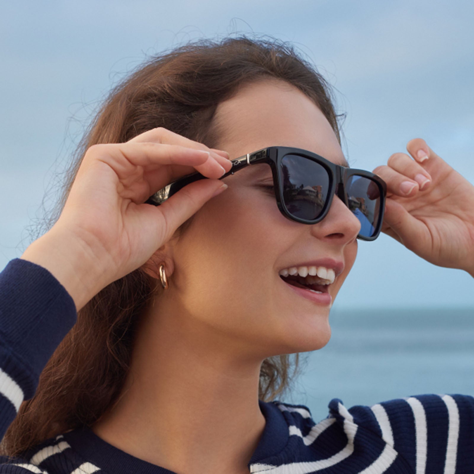 A smiling woman adjusts her Nautica Fathom black polarized wayfarer sunglasses near the ocean. She wears a navy and white striped top and gold hoop earrings, with a clear sky in the background.