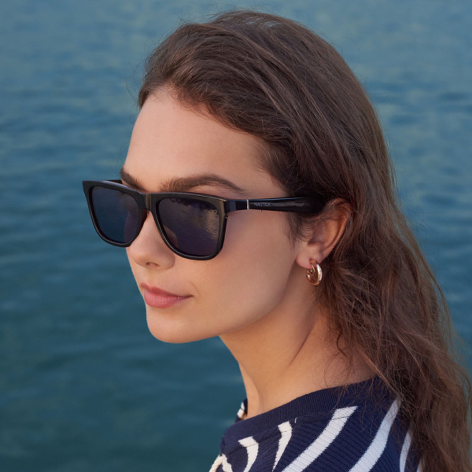 A woman with wavy brown hair and gold hoop earrings wears Nautica Fathom polarized wayfarer black sunglasses and a navy-and-white striped top, standing outdoors by calm blue water.