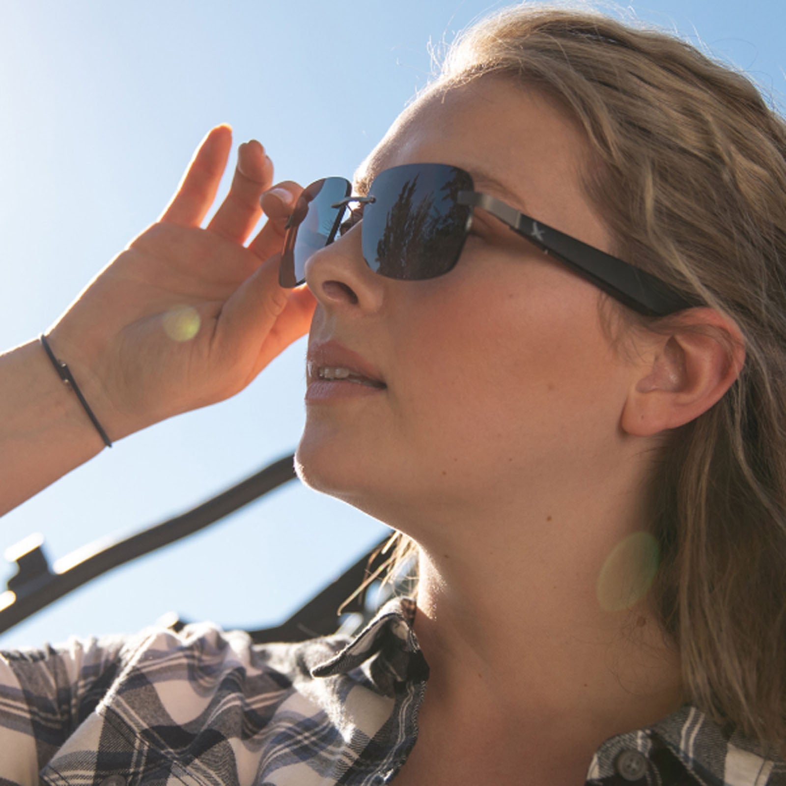 A woman in a plaid shirt adjusts her Eddie Bauer Horizon UV400 polarized sunglasses while gazing at the bright sky. Sunlight causes lens flares and highlights her blond hair on a clear day.