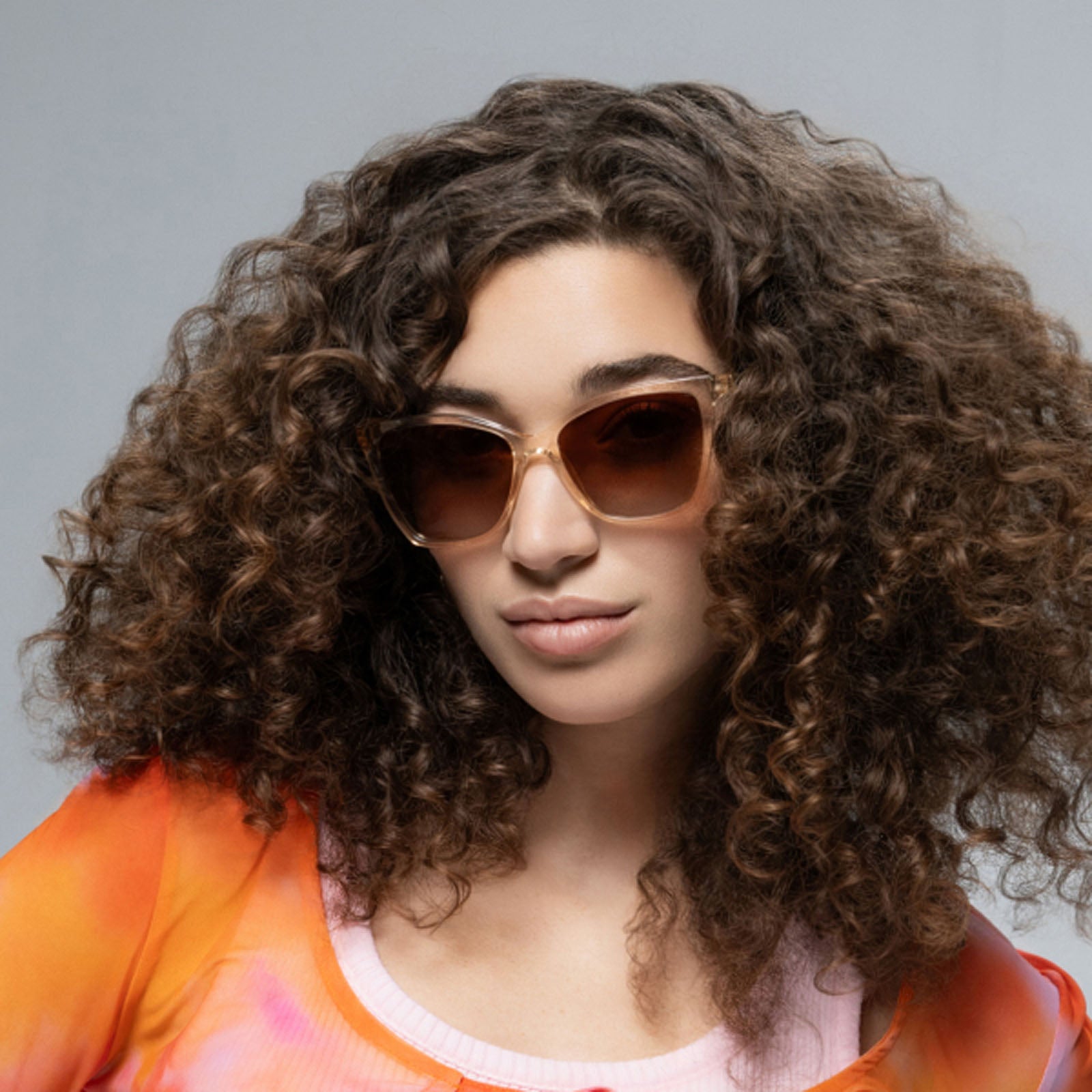 A person with voluminous curly brown hair models Lucyd Millennia women’s eyewear with brown gradient lenses and a bright orange-pink top, posing against a plain light gray background.