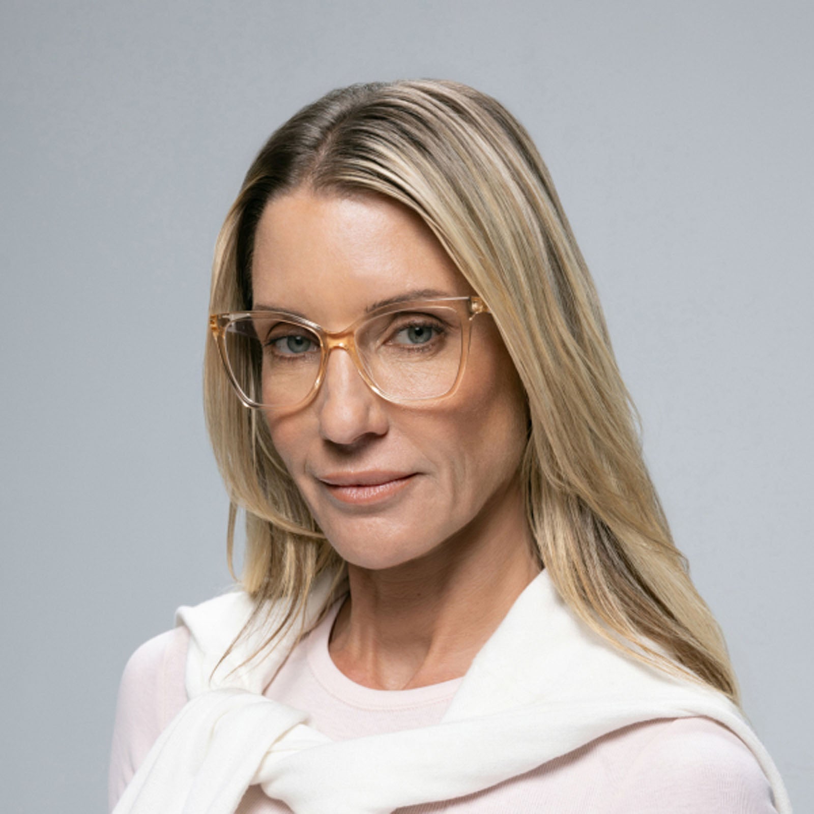 A woman with long blonde hair models Lucyd's Millennia clear-framed women's eyewear, wearing a light pink top and a white sweater draped over her shoulders, against a plain gray background.