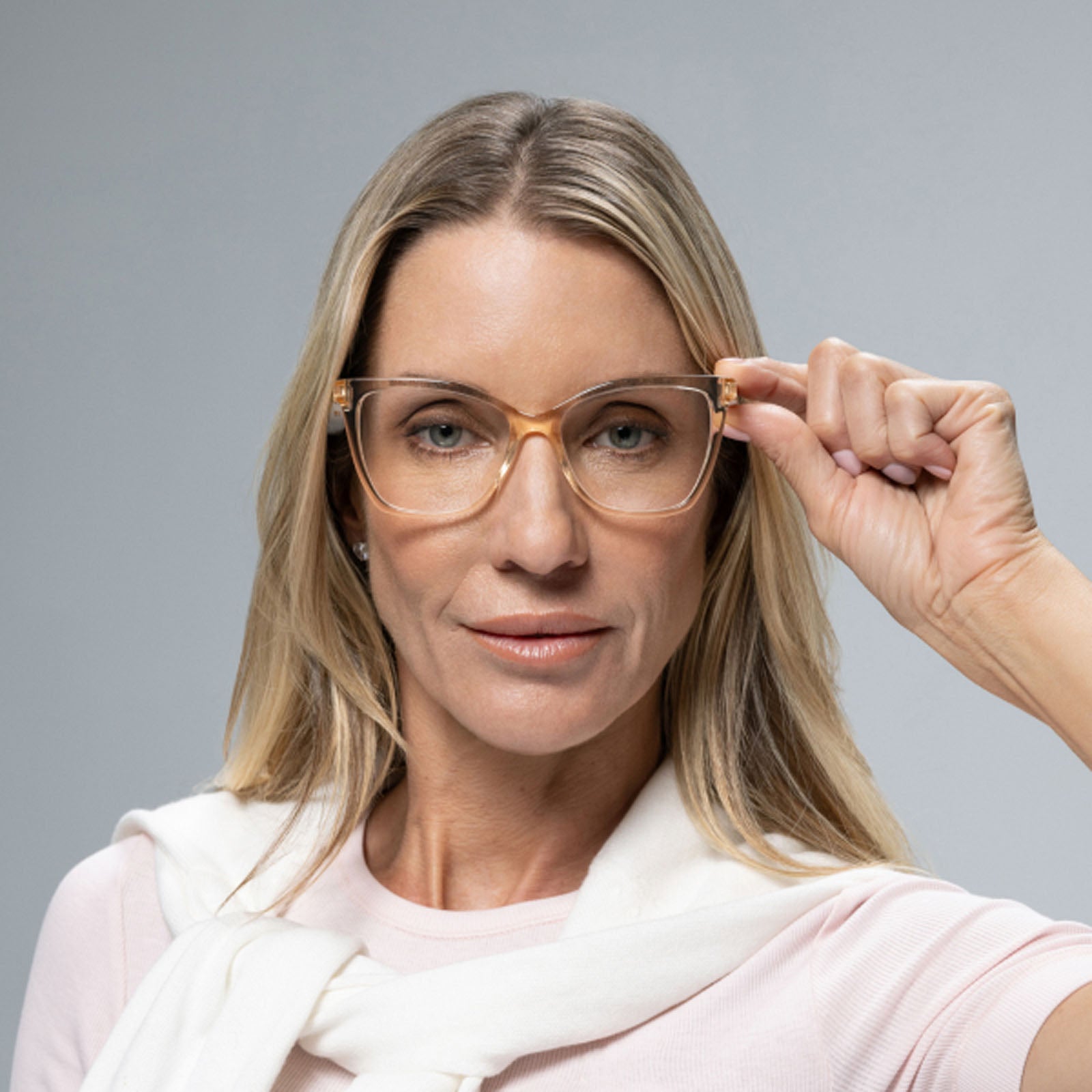 A woman with long blonde hair and light pink clothing adjusts her Lucyd Millennia brown gradient lens eyeglasses, looking confidently at the camera against a plain gray background.