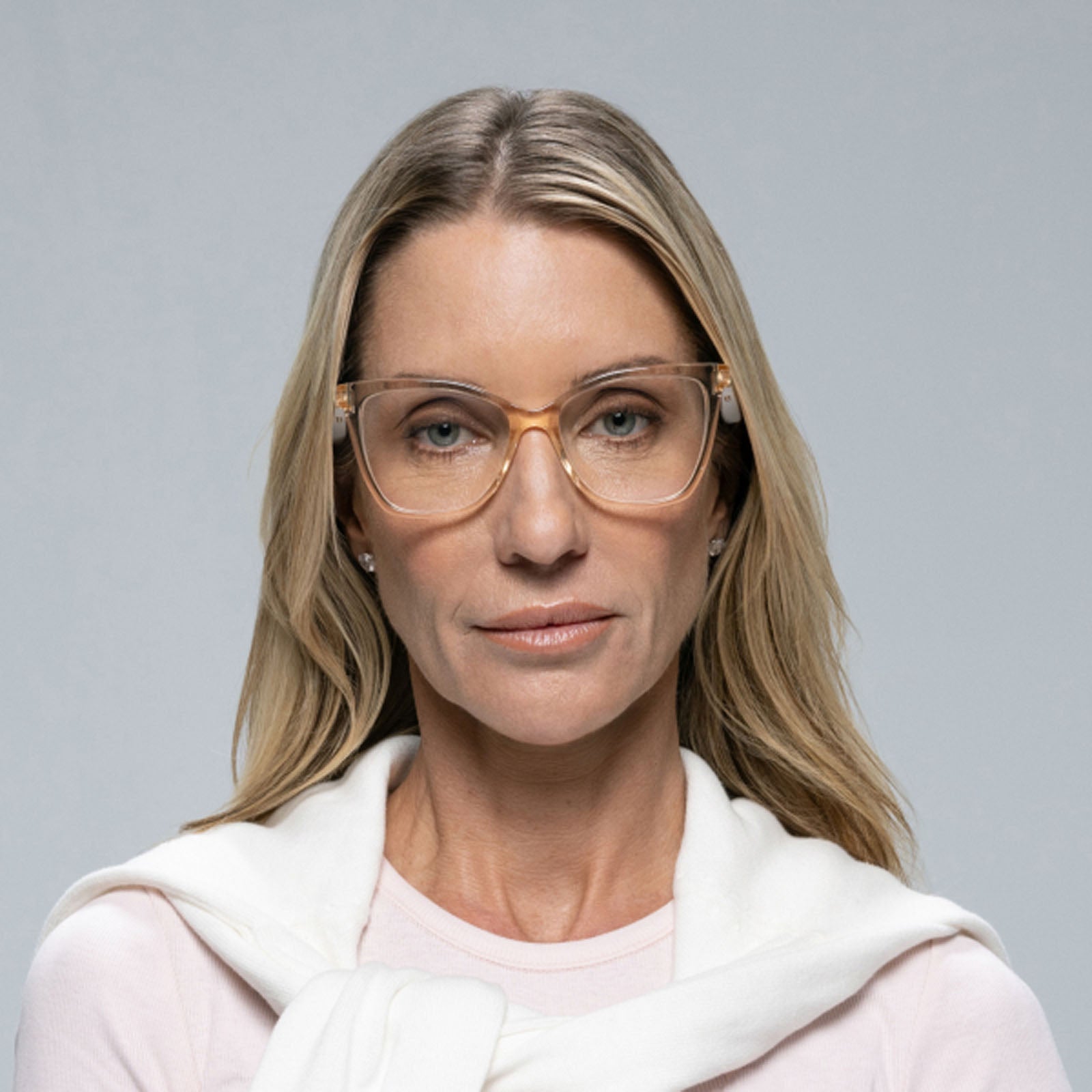A woman with long blonde hair wears Lucyd’s Millennia clear-framed eyewear, a light pink shirt, and a white sweater over her shoulders, gazing neutrally at the camera against a plain background.