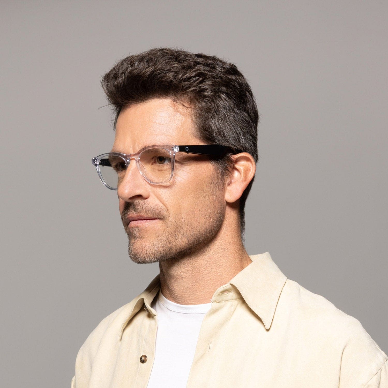 A man with short brown hair wears Lucyd Moonbeam clear-framed smart glasses and a light beige shirt over a white t-shirt, looking slightly left against a plain gray background.