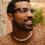 A man with short curly hair, a beard, and clear-framed Lucyd Moonbeam smart glasses is smiling and looking to the side outdoors. He wears a brown and beige patterned sweater; the background is blurred.