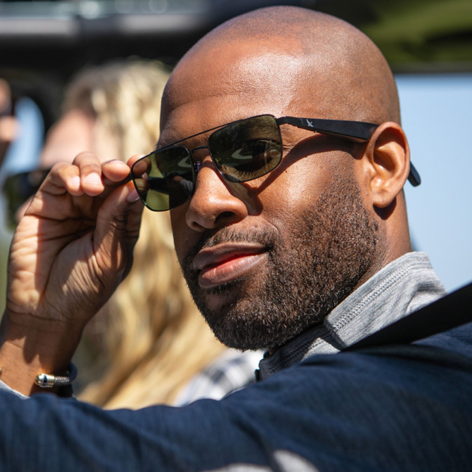 A man with a trimmed beard and shaved head smiles while adjusting his Eddie Bauer Navigator UV400 polarized sunglasses, wearing a dark jacket outdoors. The background is softly blurred, and another person can be seen.