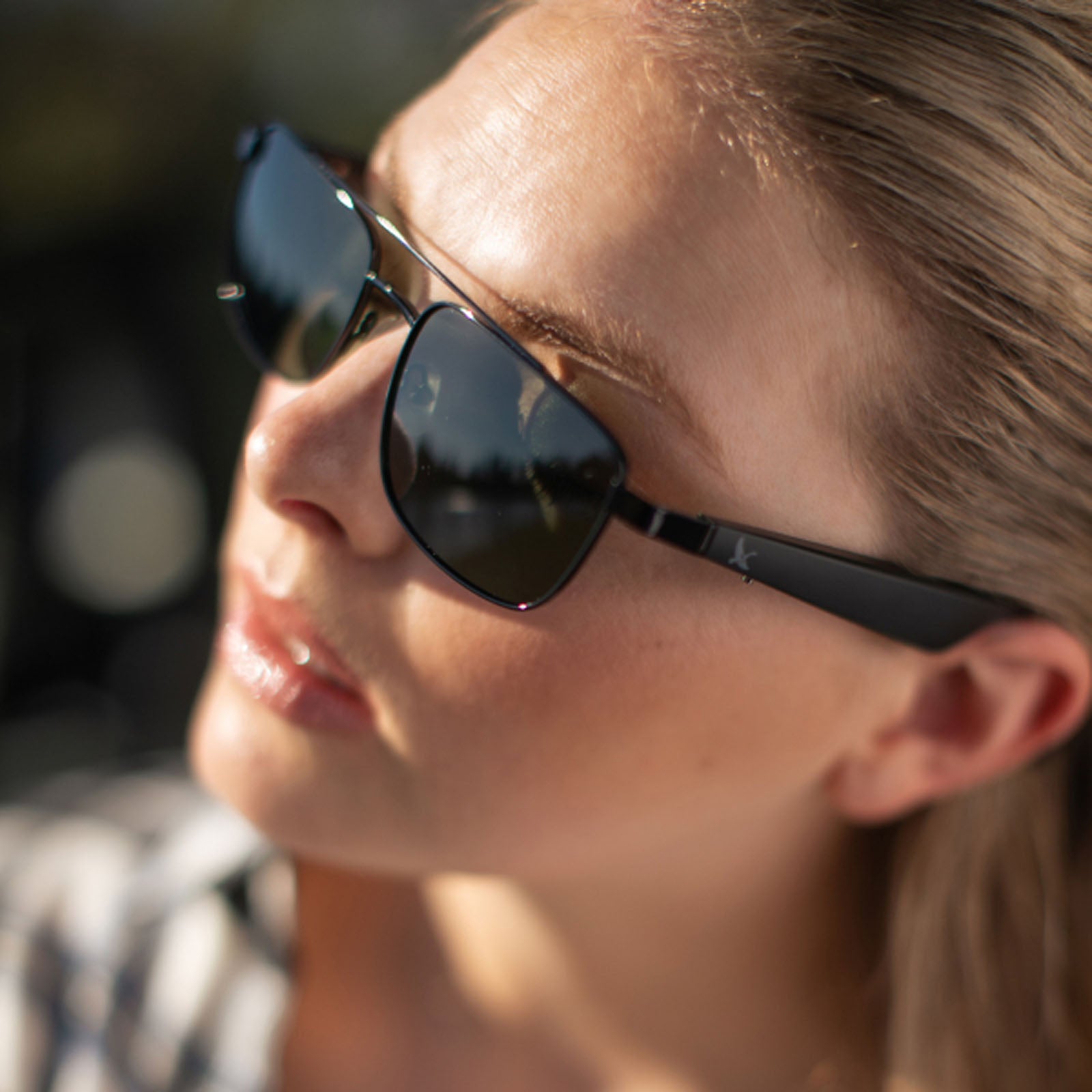 A close-up of a person with light skin and long hair wearing Eddie Bauer Navigator UV400 polarized sunglasses and a checkered shirt. Sunlight highlights their face, with the background softly blurred.