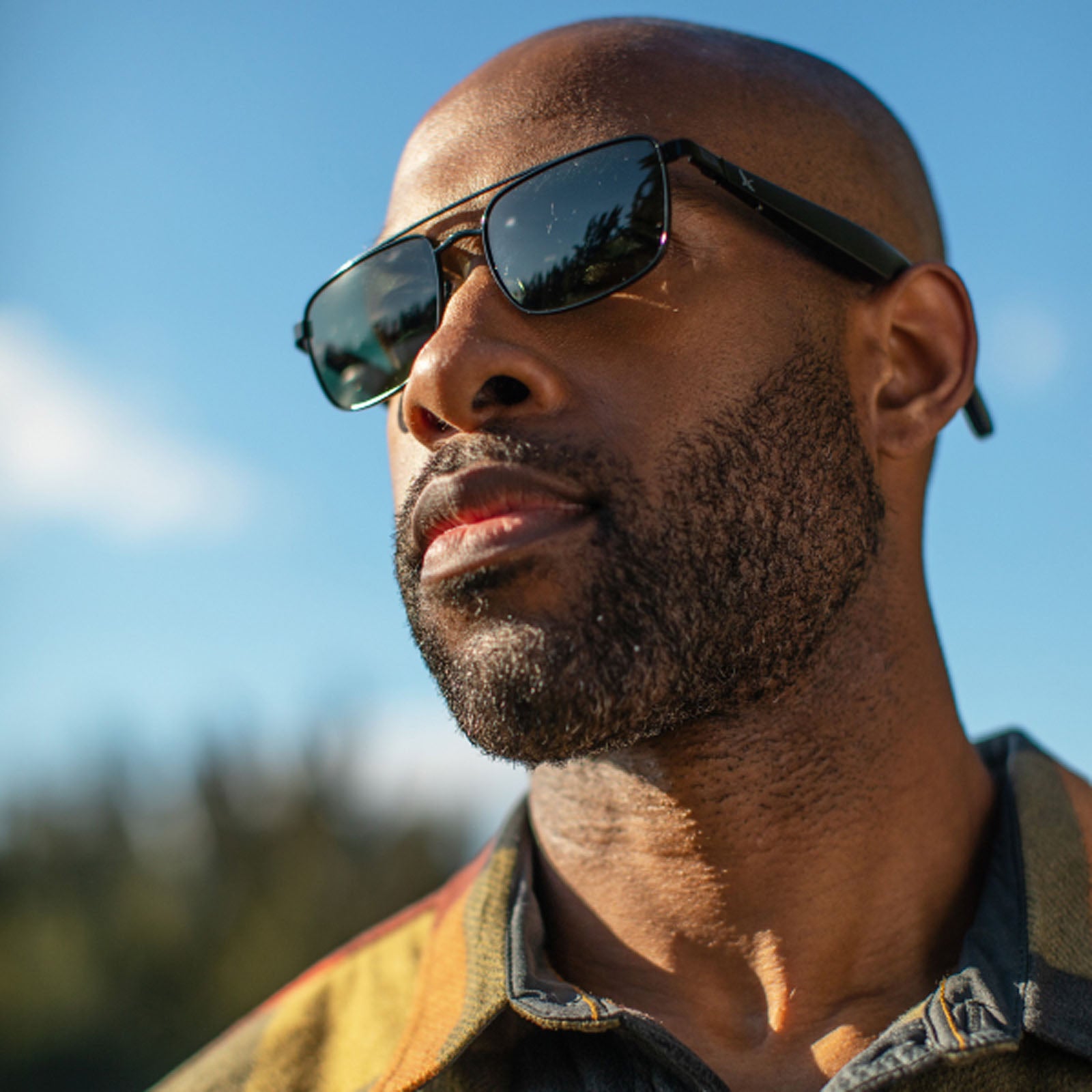 A man with a neatly trimmed beard and shaved head wears Eddie Bauer Navigator UV400 polarized sunglasses with noise-cancelling mics, gazing into the distance under a bright blue sky, with trees softly blurred in the background.