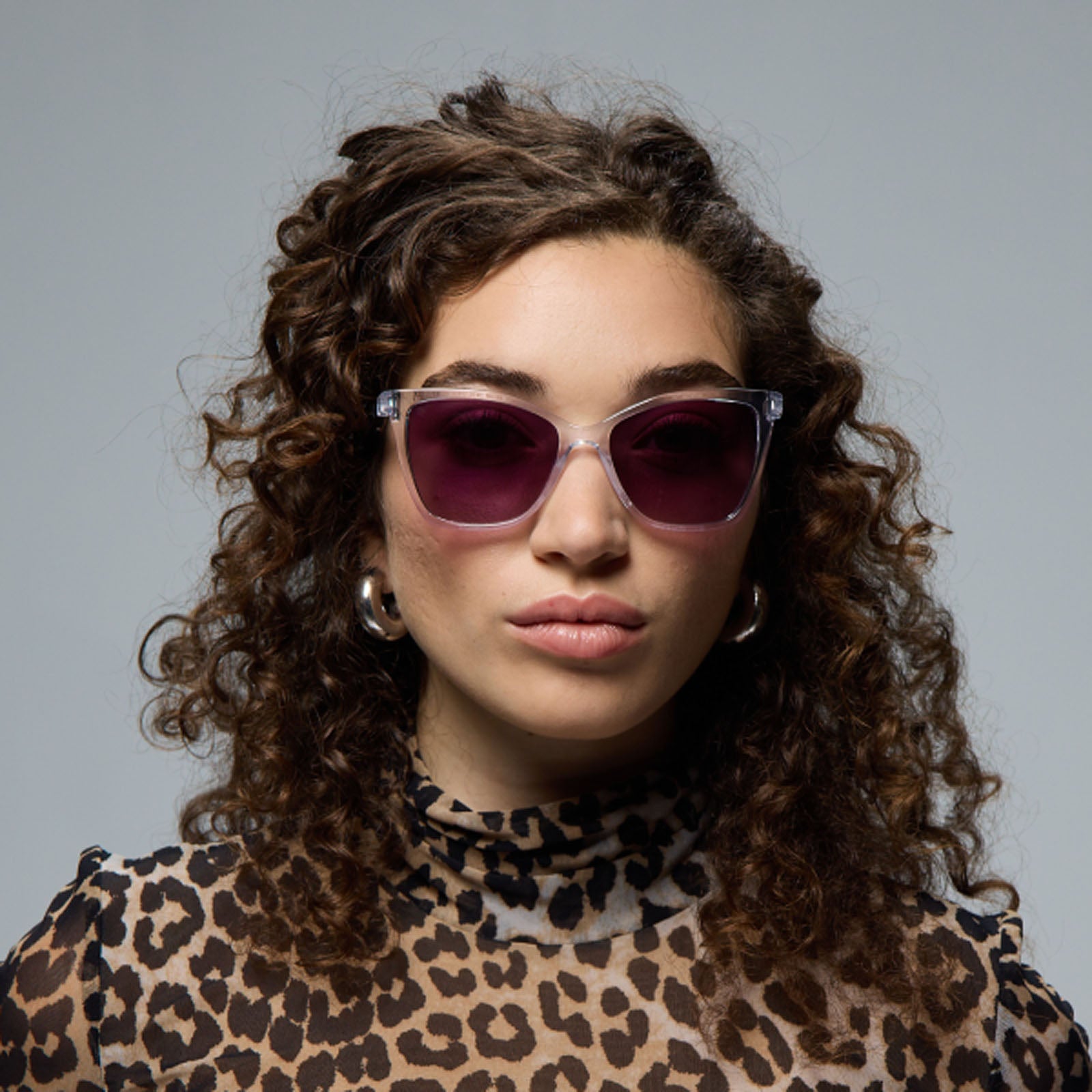 A woman with curly brown hair models Lucyd’s Moonrise women's sunglasses, featuring champagne crystal frames and polarized purple gradient lenses, paired with large hoop earrings and a leopard print top against a plain gray background.