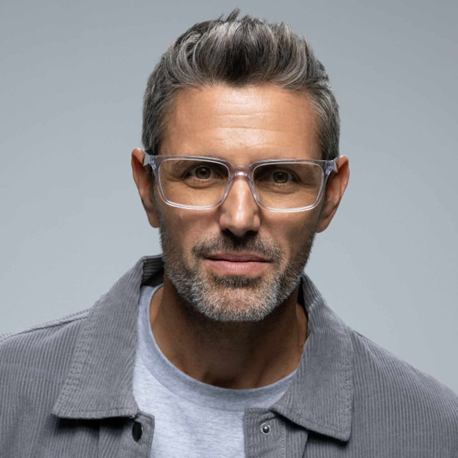A confident middle-aged man with short, gray hair and a trimmed beard wears Lucyd Eclipse smart eyewear with clear frames, paired with a gray jacket and light gray shirt, standing against a plain background.