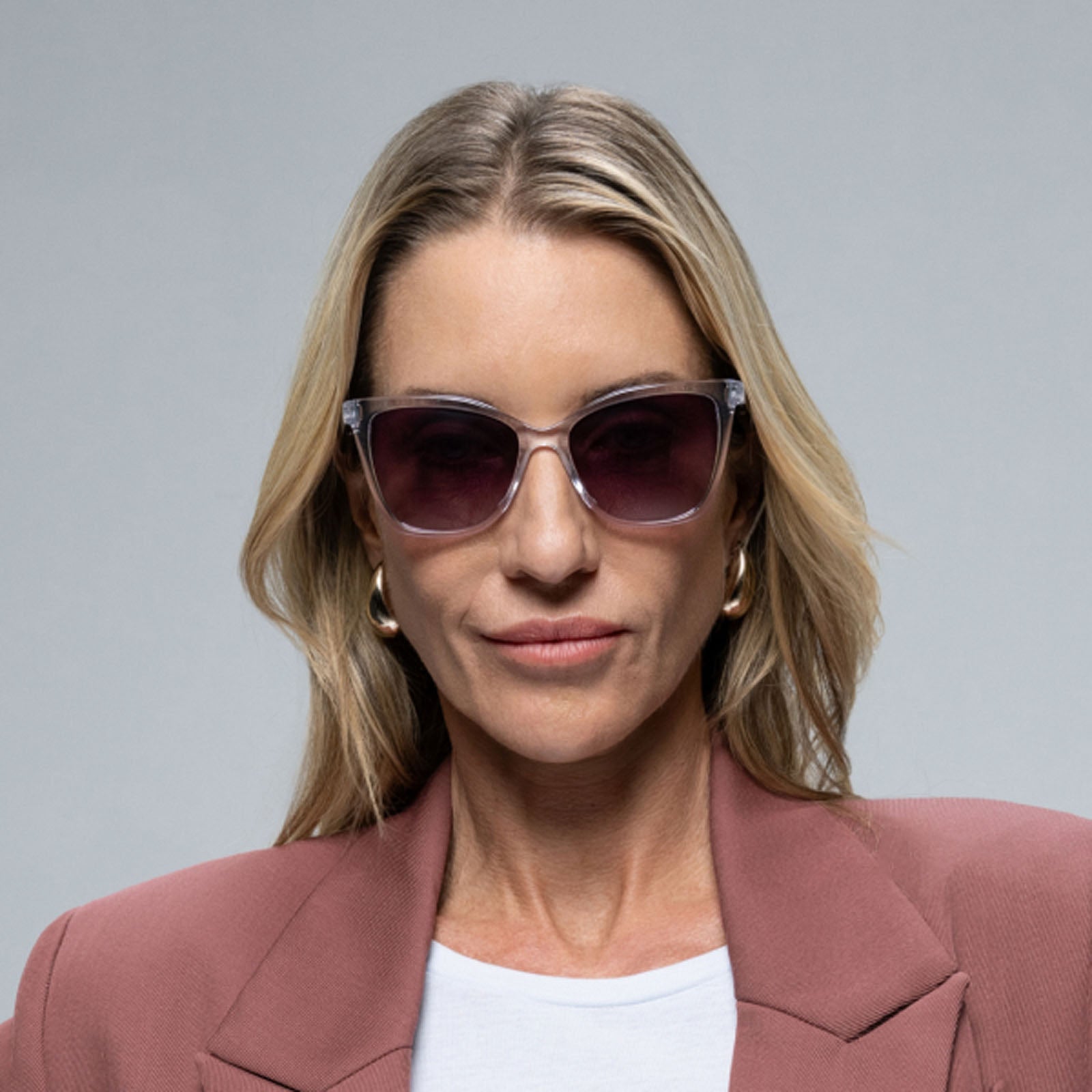A woman with long blonde hair wears Lucyd Moonrise champagne crystal sunglasses with polarized purple gradient lenses, a mauve blazer, white top, and gold hoop earrings against a plain gray background.