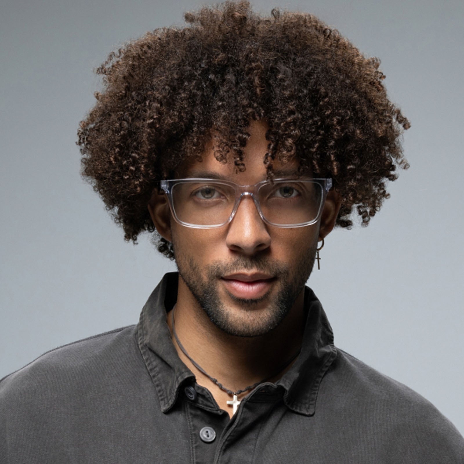 A man with curly hair models the Lucyd Eclipse smart eyewear, paired with a dark button-up shirt, cross necklaces, and a small hoop earring against a plain gray background.
