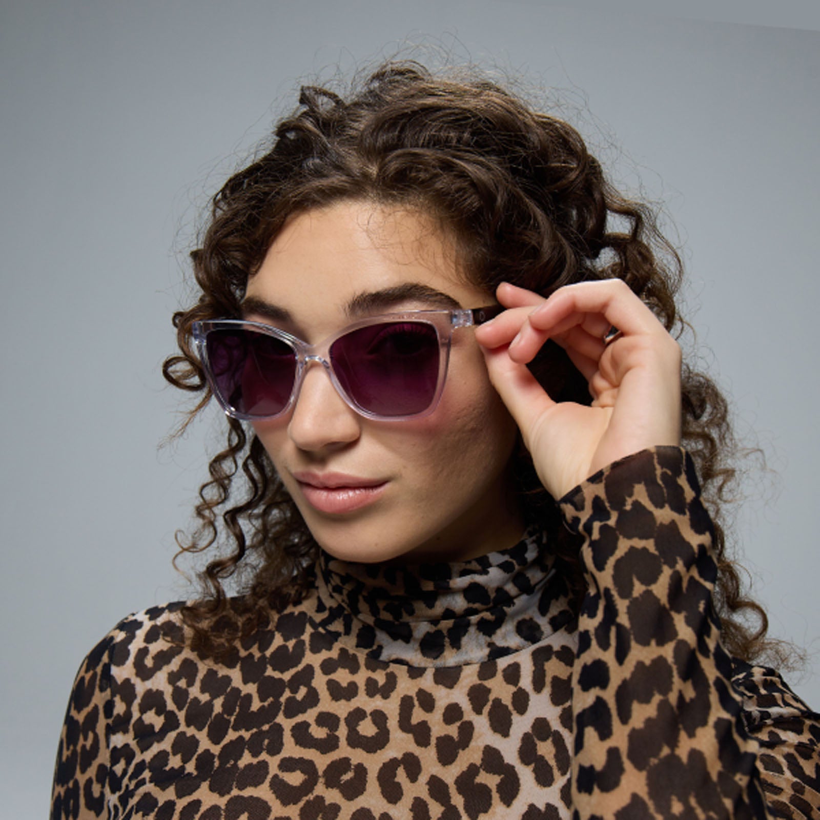 A woman with curly hair in a leopard print turtleneck confidently touches the frame of her Lucyd Moonrise sunglasses, featuring polarized purple gradient lenses.