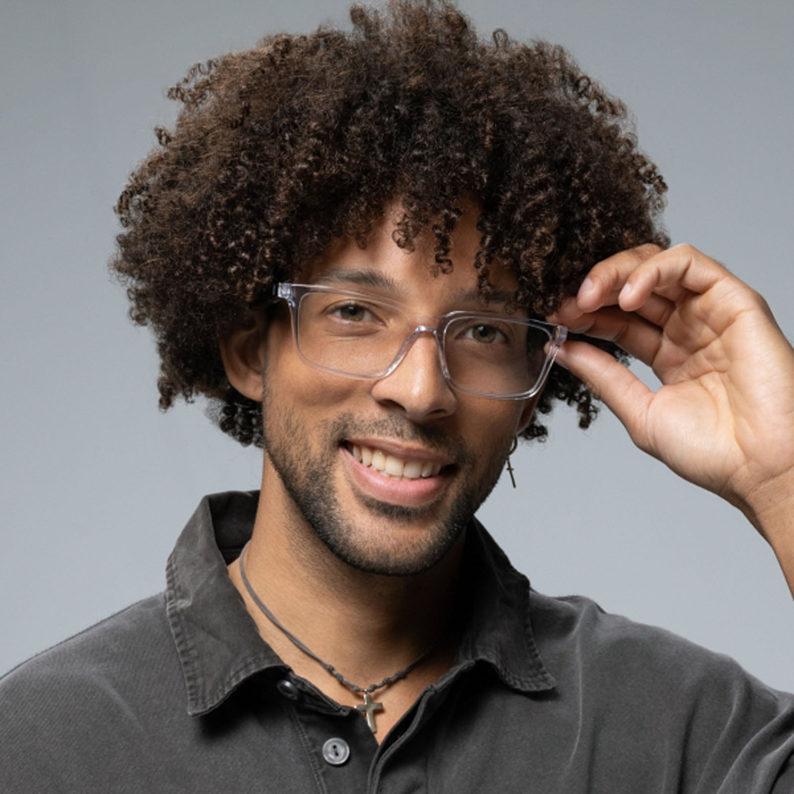 A smiling man with curly hair and a short beard adjusts his Lucyd Eclipse smart eyewear with clear frames, wearing a dark collared shirt and necklace against a plain gray background.