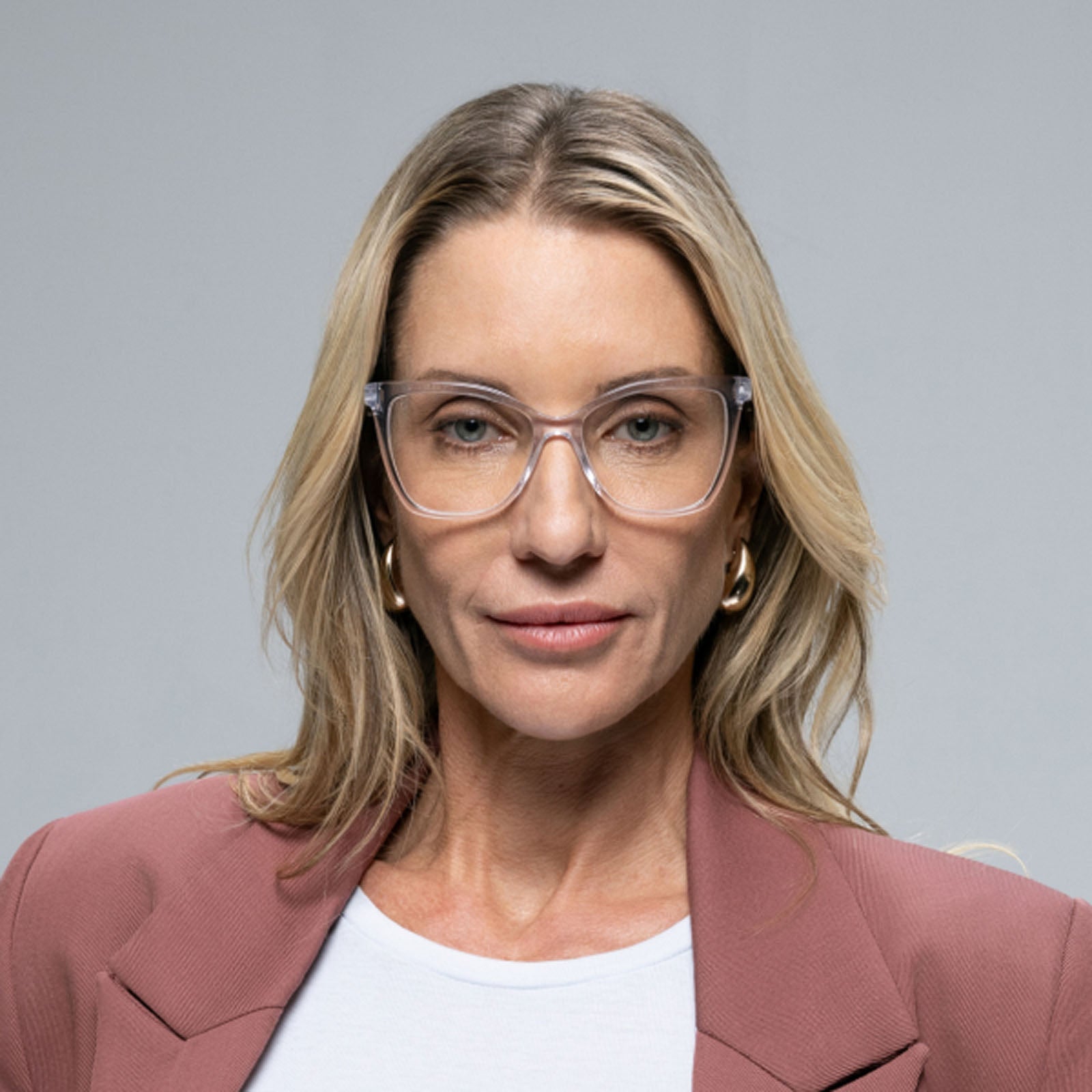 A woman with long blonde hair wears Lucyd Moonrise champagne crystal sunglasses, gold hoop earrings, a white top, and a mauve blazer while posing against a plain gray background.