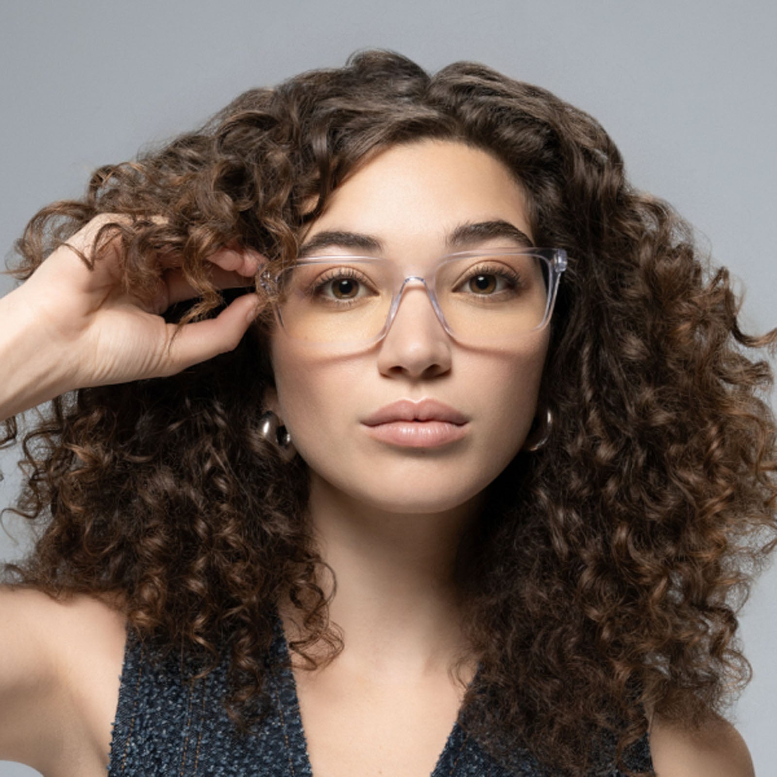 A person with long, curly brown hair and light skin wears Lucyd Eclipse clear-framed smart eyewear and silver hoop earrings, looking at the camera and touching their glasses against a plain gray background.