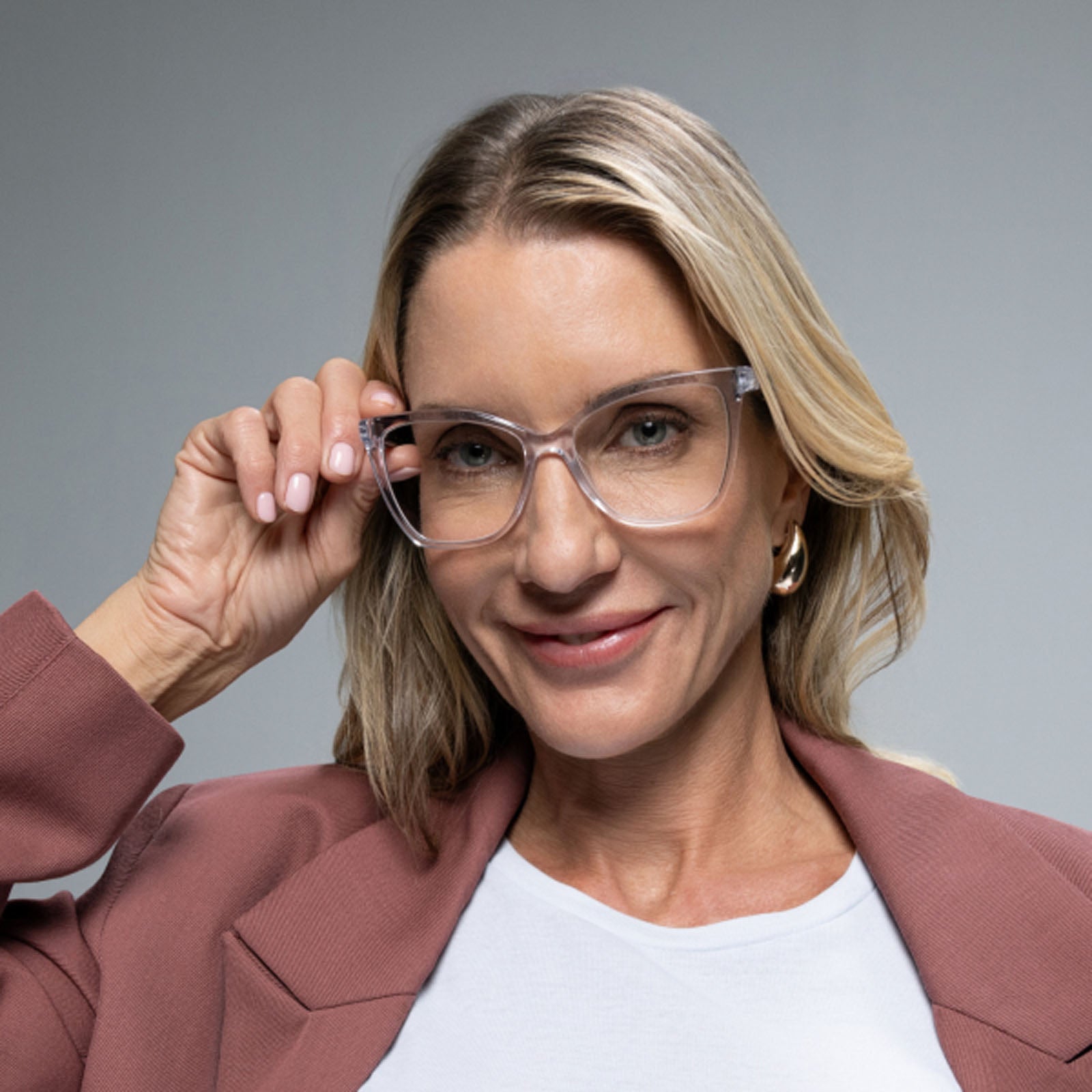 A blonde woman smiles against a plain gray background, wearing Lucyd’s Moonrise champagne crystal sunglasses and a mauve blazer over a white top, holding her glasses with one hand.
